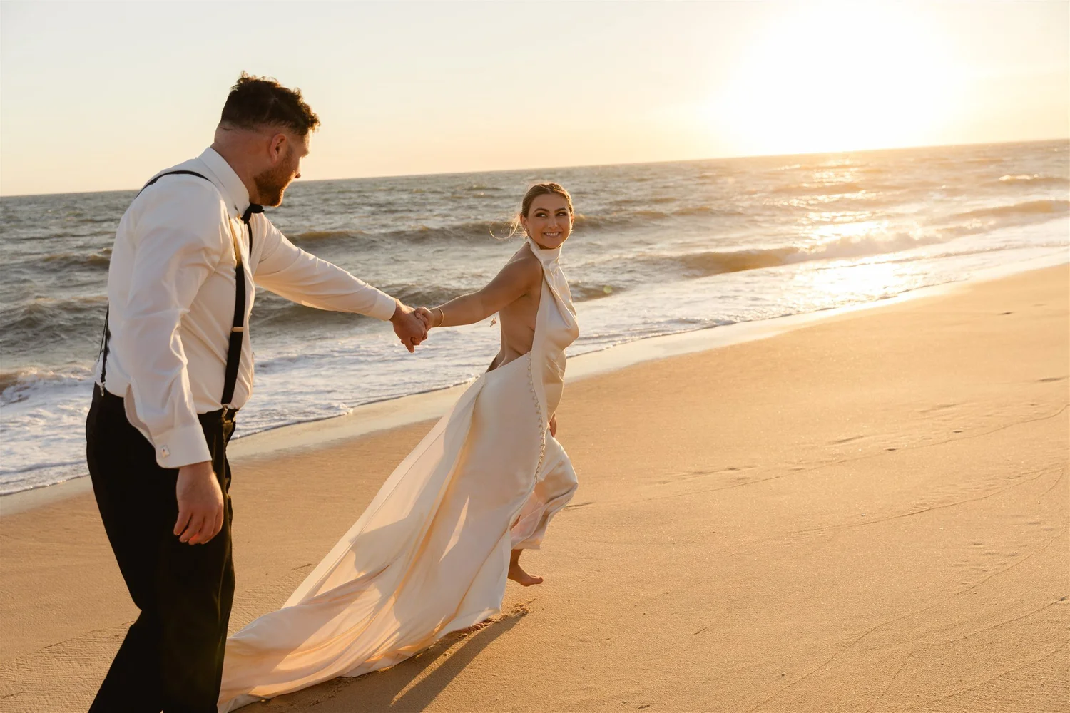 A couple holding hands on the beach during sunset, the woman in a white wedding dress and the man in formal attire.