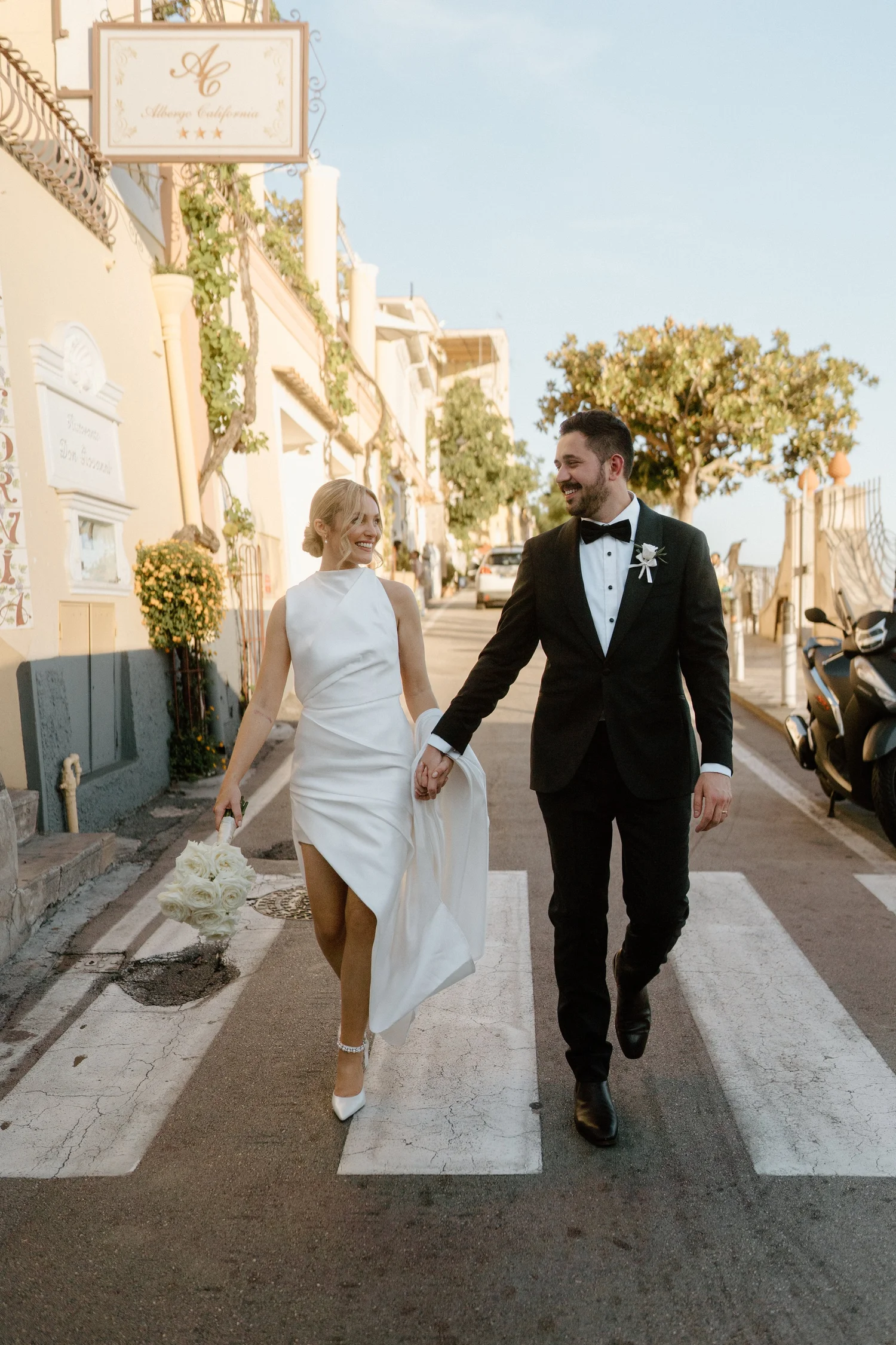 A newlywed couple walking hand in hand on a city street during daytime, with the bride holding a bouquet of white roses and smiling at the groom, who is dressed in a black tuxedo with a bow tie and a boutonniere, in front of a colorful building with
