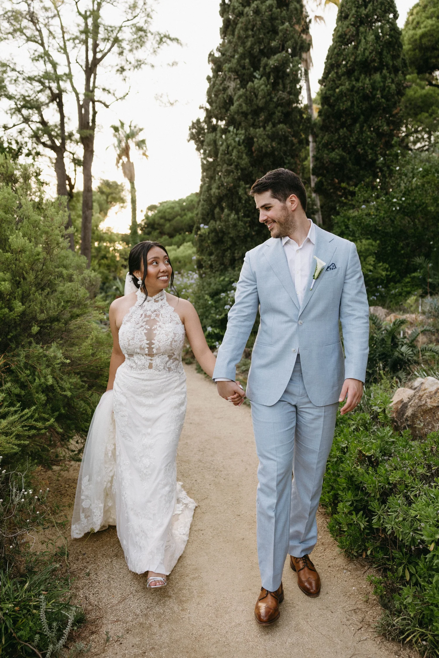 A newlywed couple walks hand-in-hand along a garden path, surrounded by lush greenery and tall trees. The bride is wearing a sleeveless white lace wedding dress and heels, and the groom is dressed in a light blue suit with brown shoes. They are smili