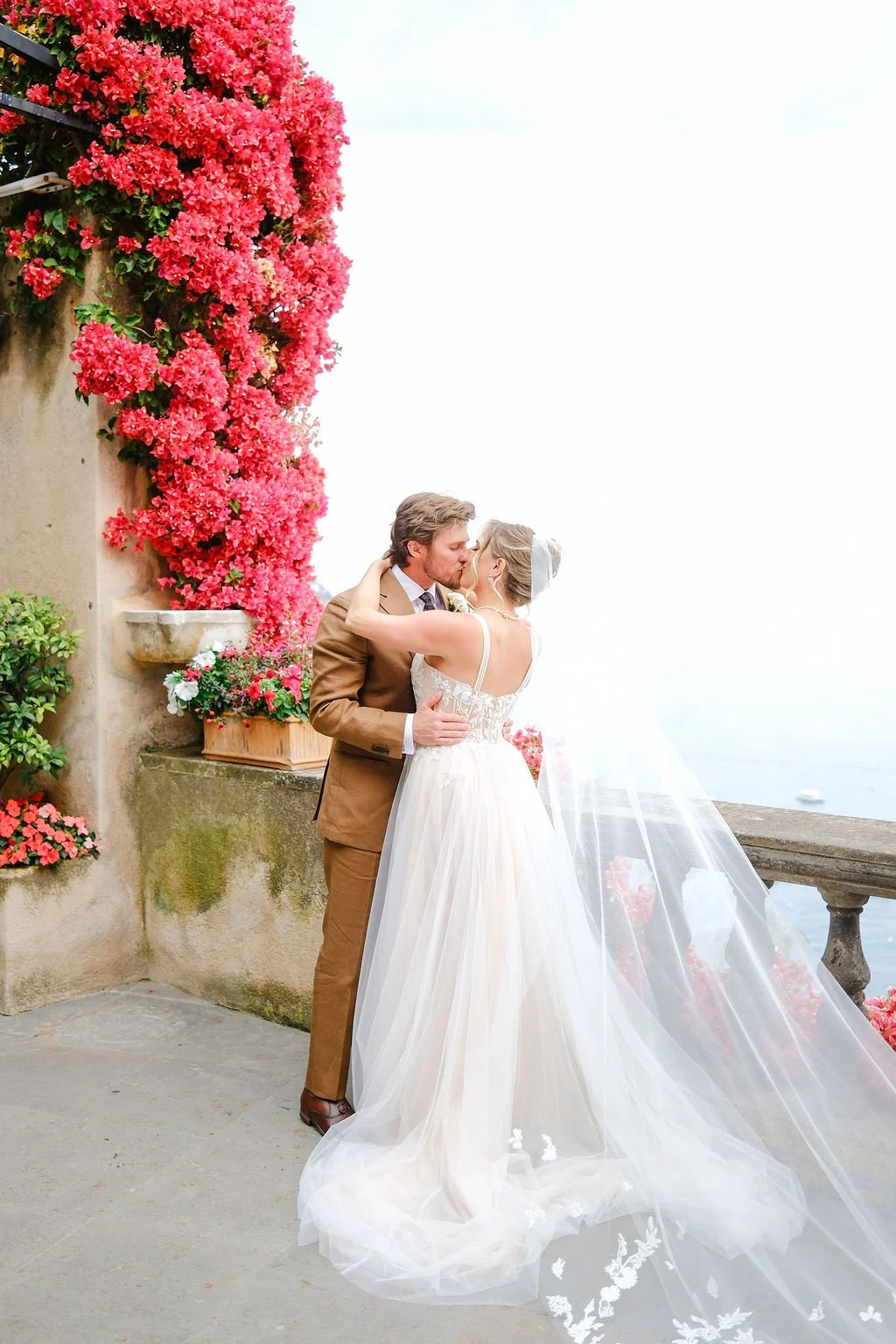 A bride and groom share a kiss on a balcony decorated with pink flowers, overlooking the water.