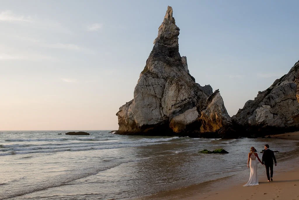 A couple dressed in wedding attire walking hand in hand on a beach with a large rock formation in the background during sunset.