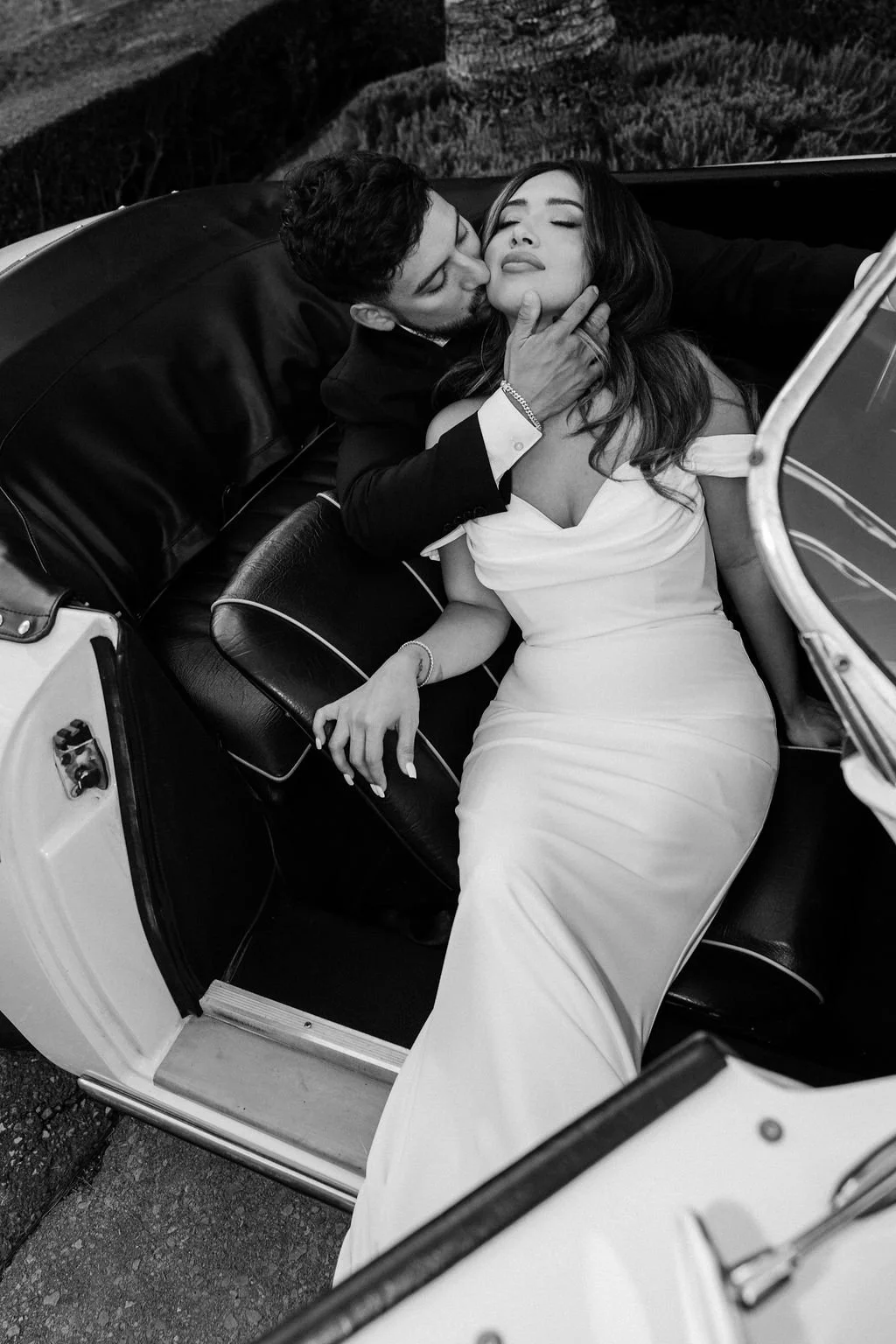 A man and woman sitting in a vintage convertible car, with the man kissing the woman's cheek and holding her chin, while the woman has her eyes closed and appears to enjoy the moment. The woman is dressed in an elegant off-shoulder dress, and the sce