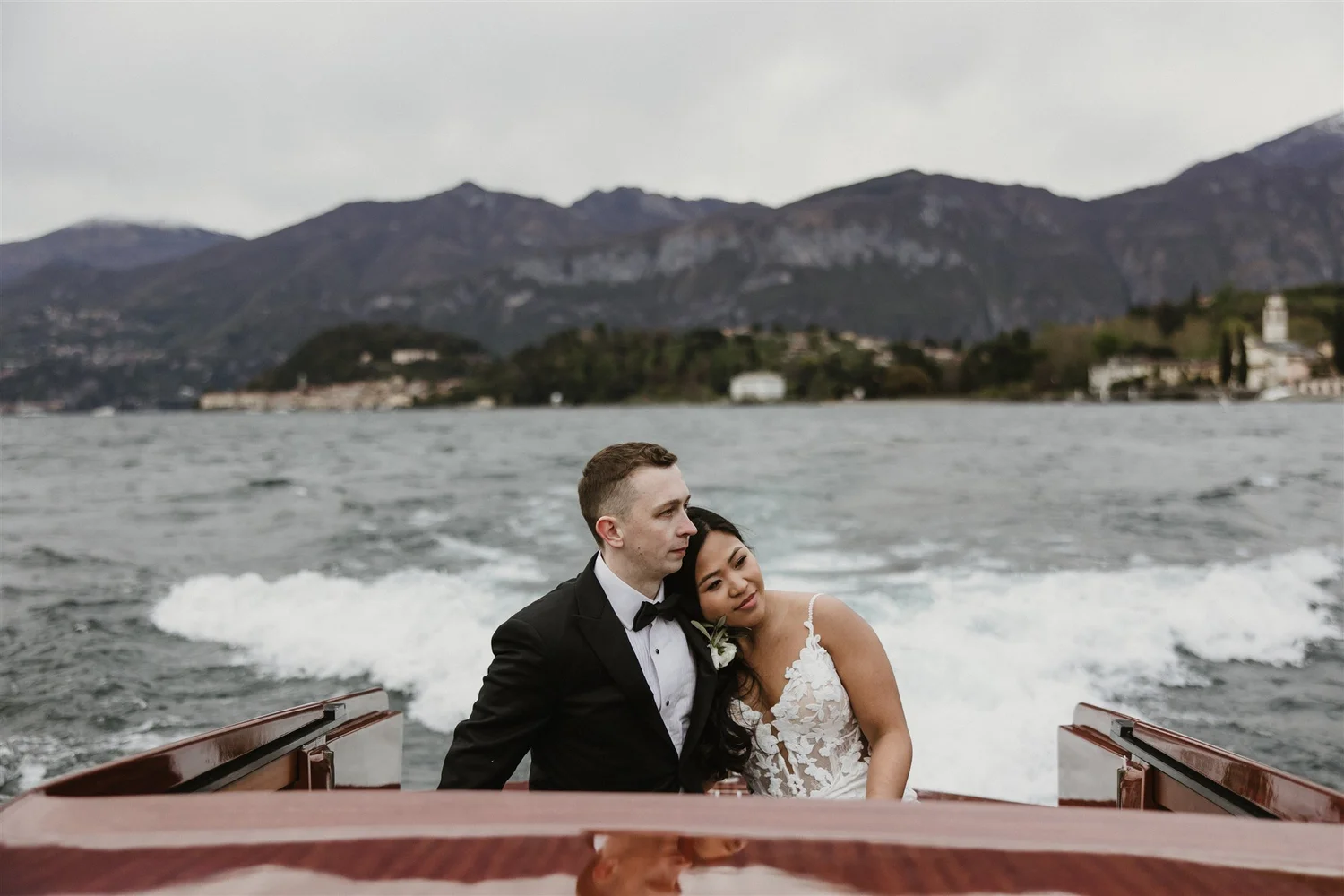 A newlywed couple dressed in wedding attire sitting together on a boat on a lake with mountains and a cloudy sky in the background.