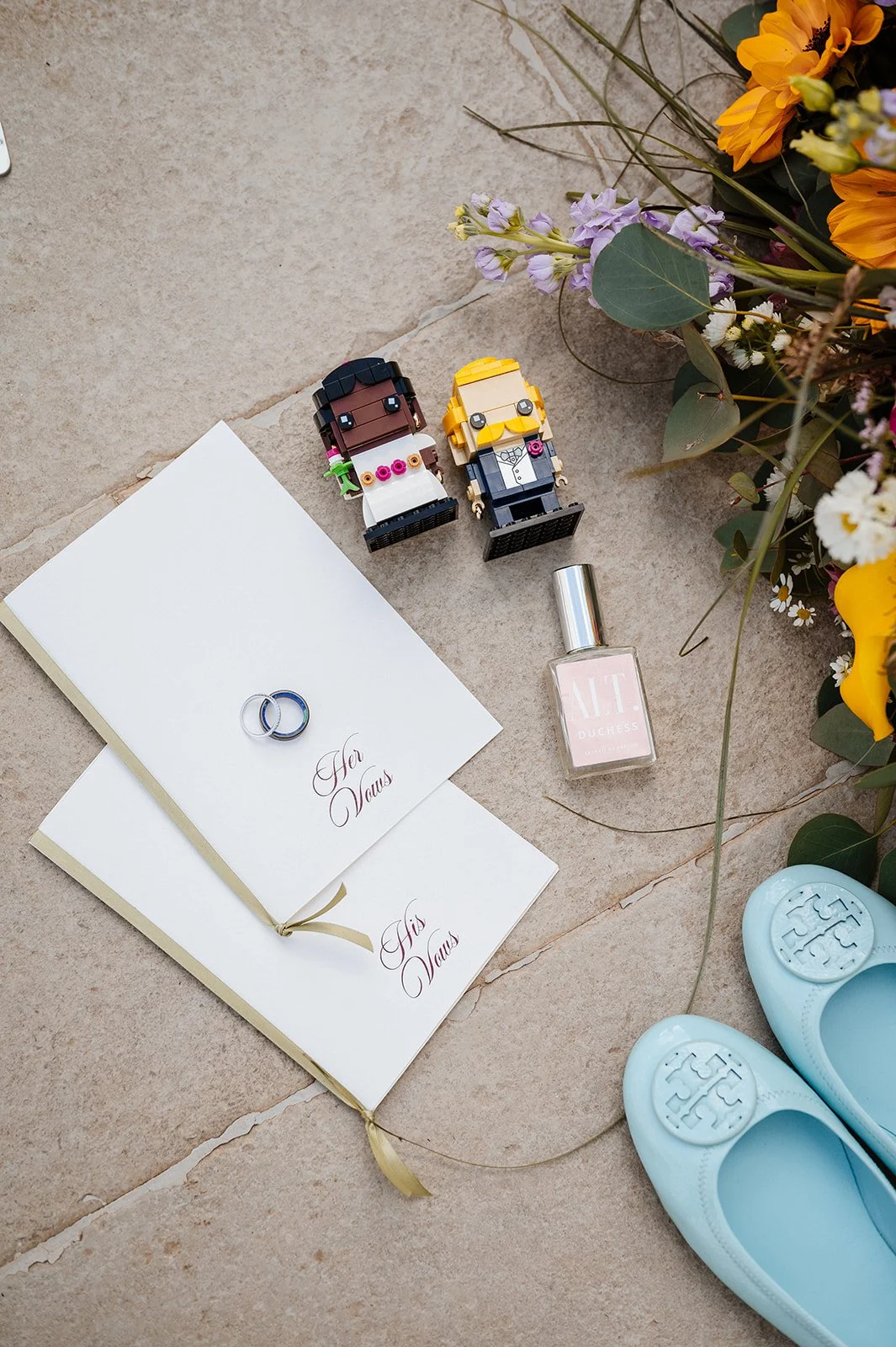 A flat lay of wedding or engagement items including two open notebooks with gold ribbons, two small LEGO bride and groom figures, a bottle of perfume labeled 'Duchess,' a pair of light blue TOMS shoes, and a bouquet of assorted flowers on a beige til