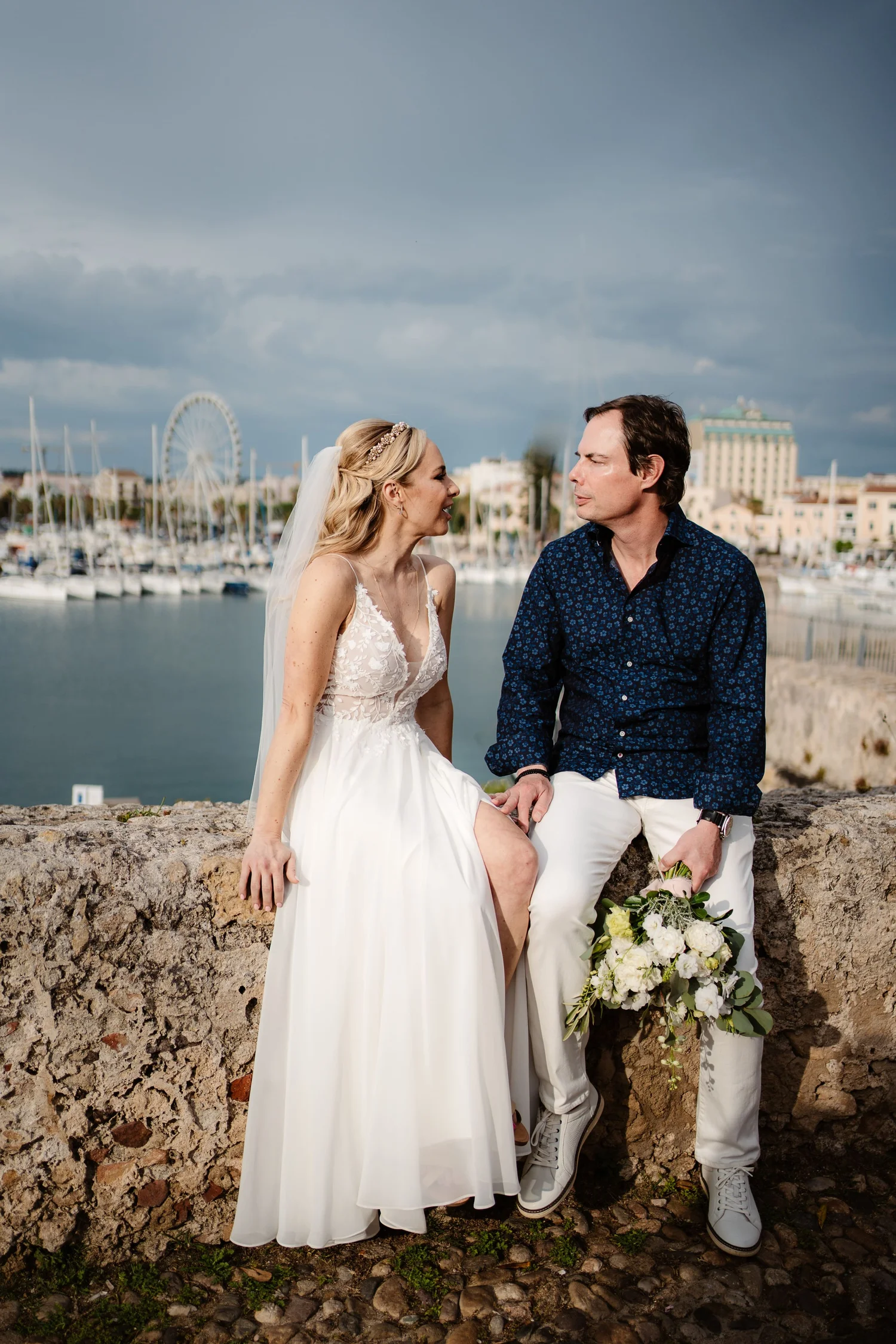 A bride and groom sit on a stone wall by a marina with sailboats, a Ferris wheel, and city buildings in the background, sharing an intimate moment.