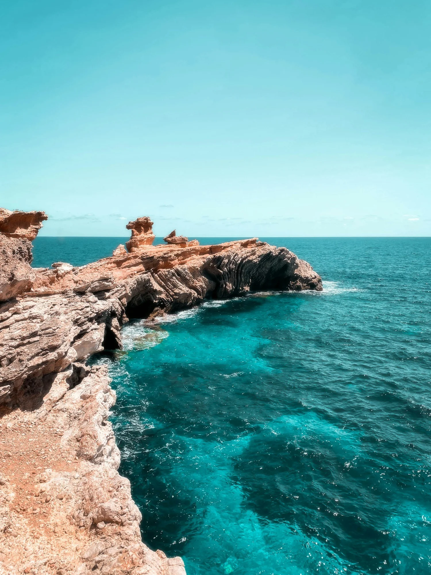 Scenic view of rugged red rock cliffs over a blue ocean with a light blue sky.