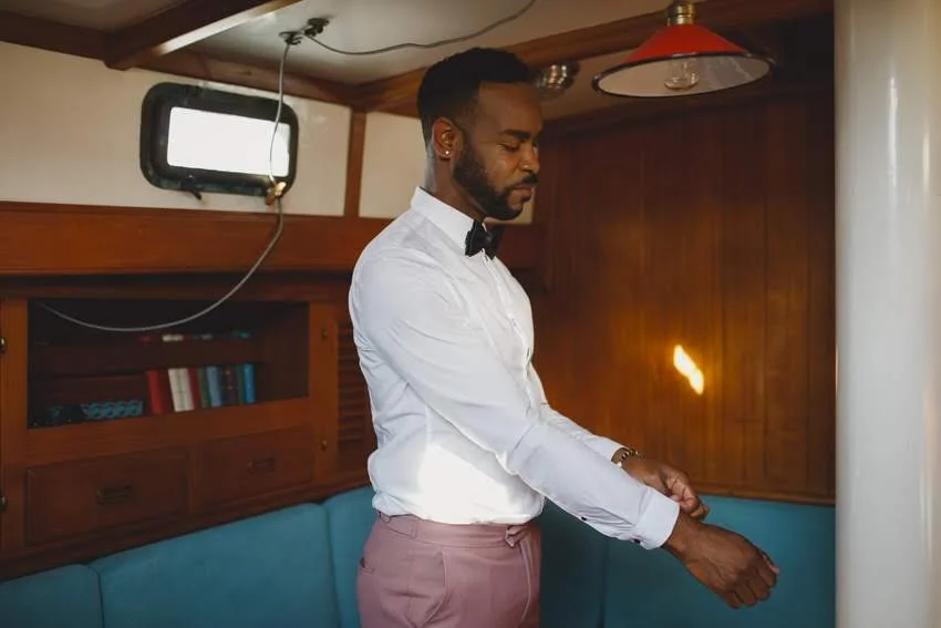 A man in a white shirt and pink pants adjusting his sleeves inside a wooden-paneled room with blue seating.