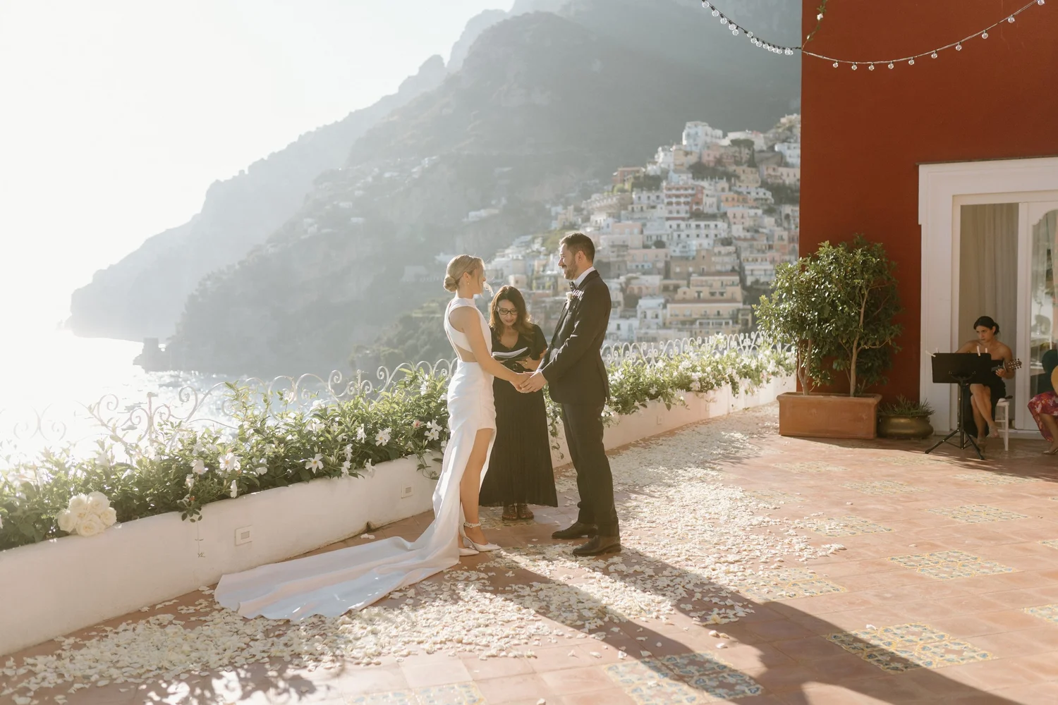A couple getting married outdoors on a terrace with a view of a hillside town, with a musician playing guitar in the background and a woman officiating.