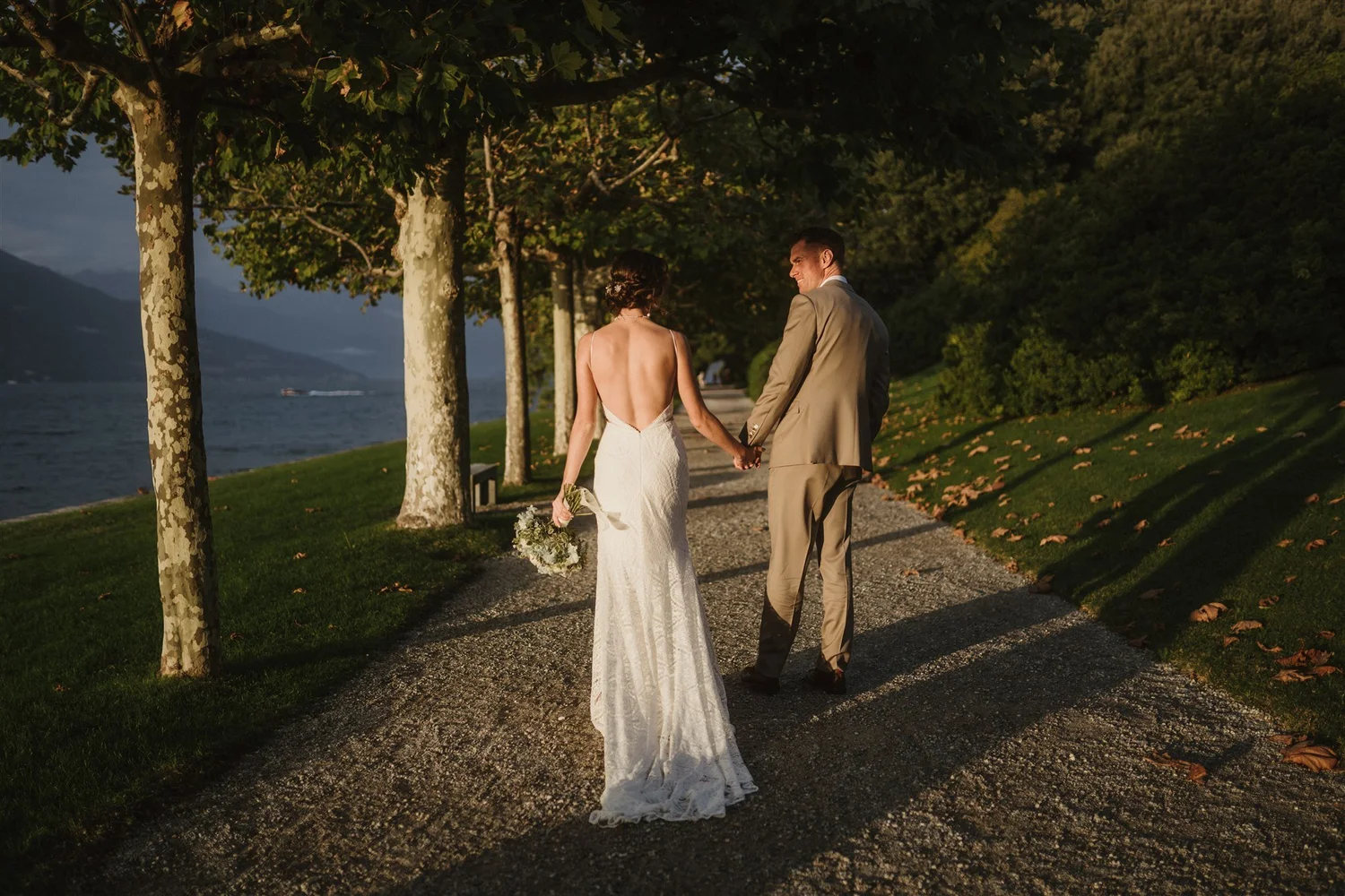 A bride and groom holding hands walking under a tree-lined path overlooking a body of water during sunset.