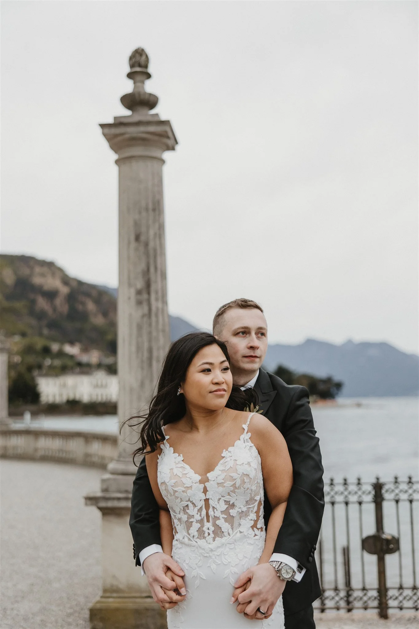 A bride and groom standing close together outdoors near a lake, with a stone pillar and mountains in the background. The bride is wearing a white lace wedding dress, and the groom is in a black tuxedo. They are embracing with the groom's arms around 