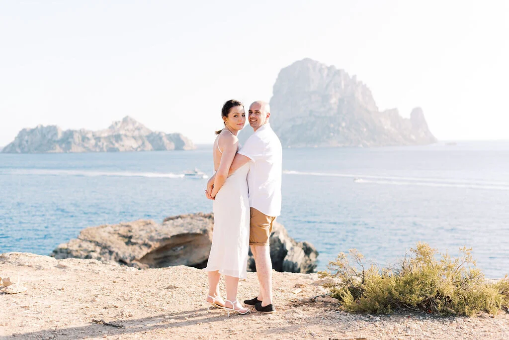 A couple stands embracing on a rocky shoreline with the ocean and large island in the background, during daytime.