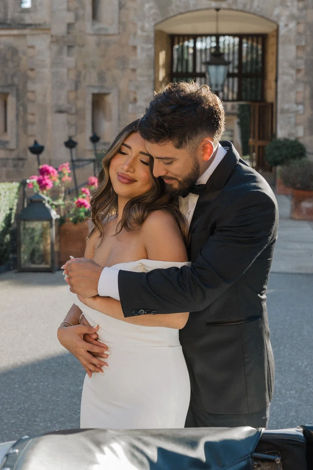 A bride and groom hugging outside a stone building, with the bride smiling and the groom resting his face on her head.