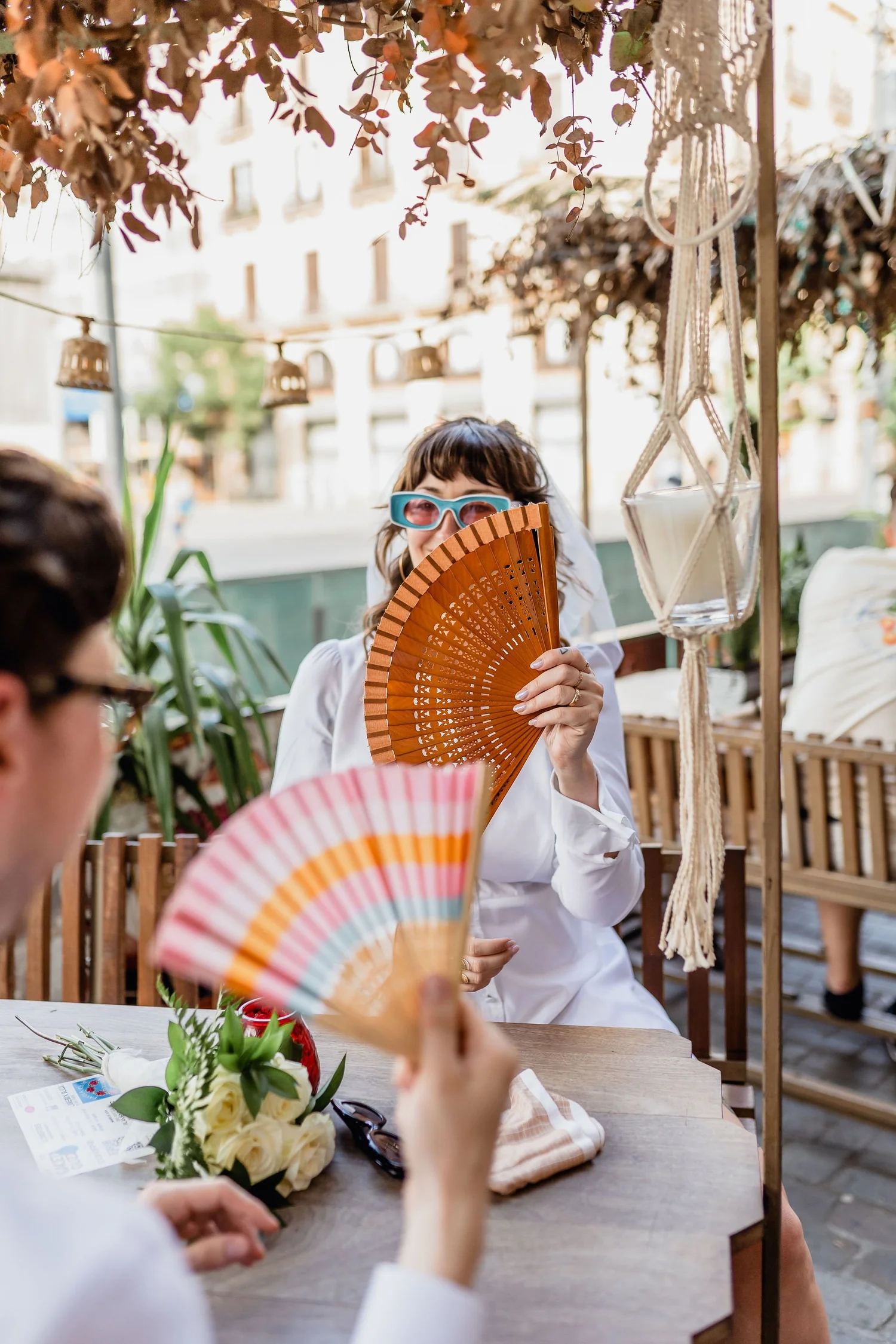 A woman with short brown hair, wearing sunglasses and a white outfit, is holding a brown fan covering part of her face. She is seated at an outdoor table decorated with a bouquet of white flowers and greenery. The setting appears to be a sunny outdoo