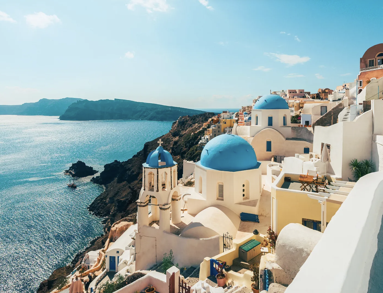 Scenic view of Santorini, Greece, with white buildings and blue-domed roofs overlooking the Aegean Sea on a sunny day.