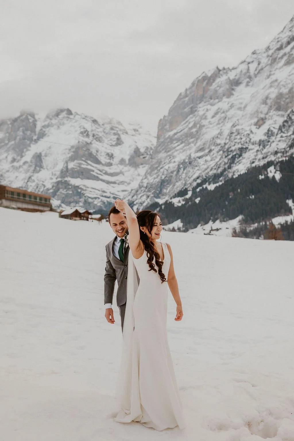 A smiling couple in wedding attire walking in the snow with snow-covered mountains in the background.