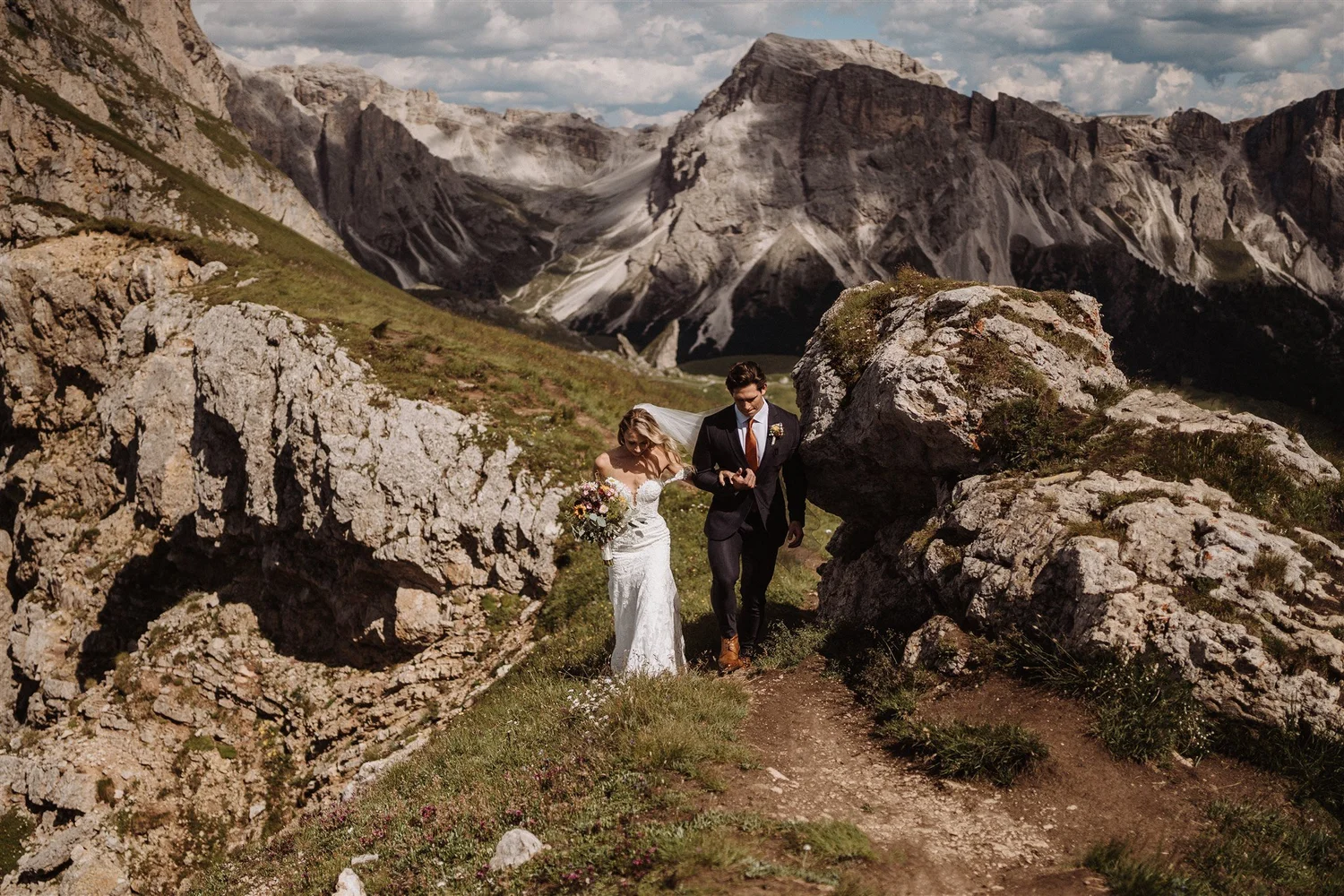 A bride and groom walking through rocky terrain with mountain peaks and partly cloudy sky in the background.