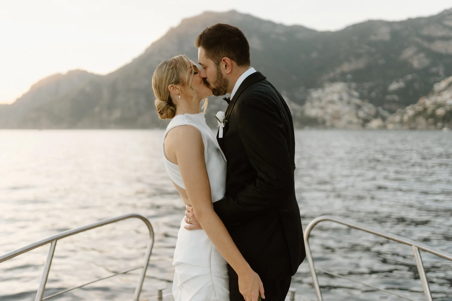 A bride and groom kissing on a boat with mountains and water in the background, during sunset.
