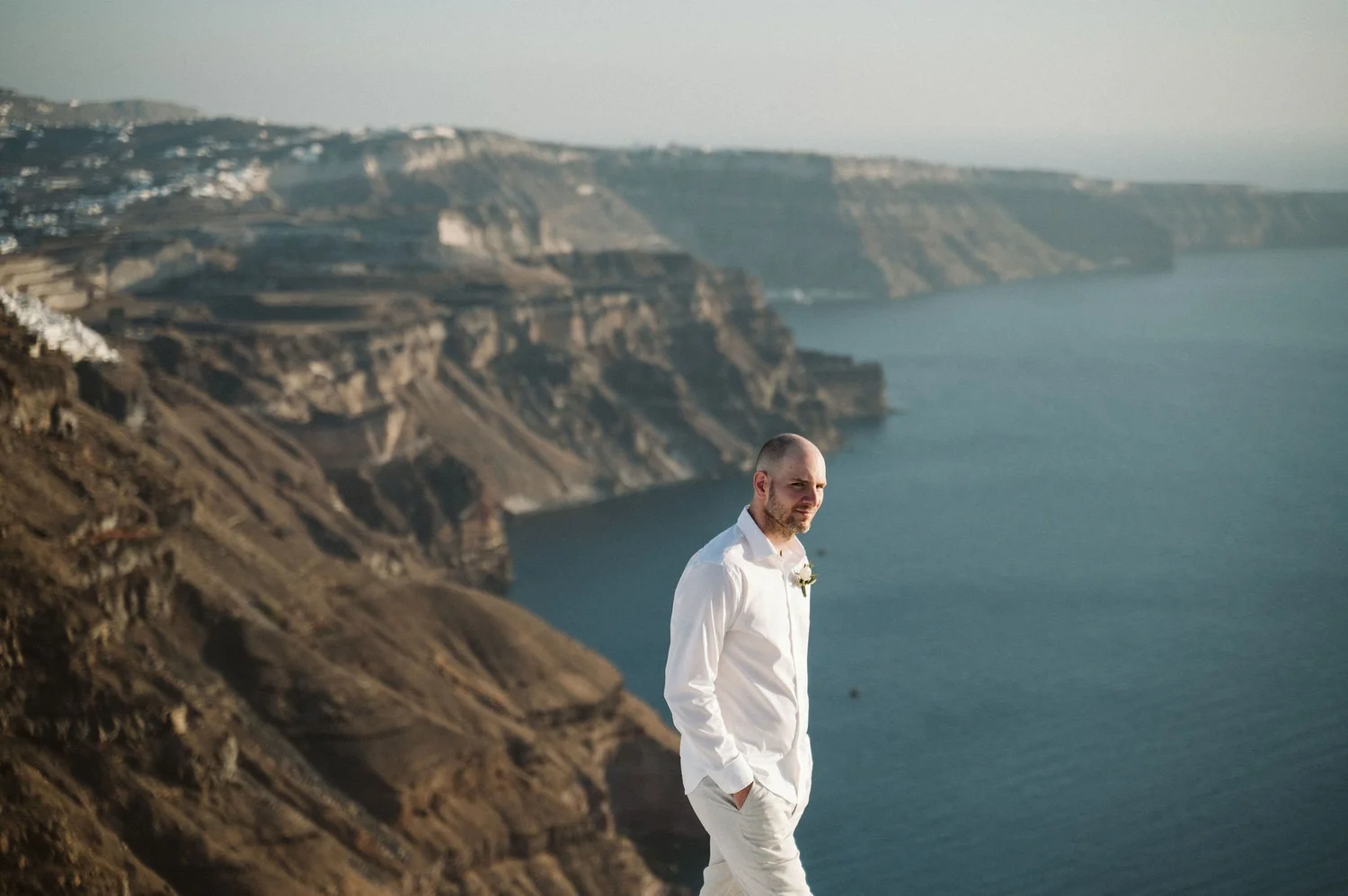 A man in a white shirt and white pants standing on a cliffside overlooking the ocean with cliffs in the background.