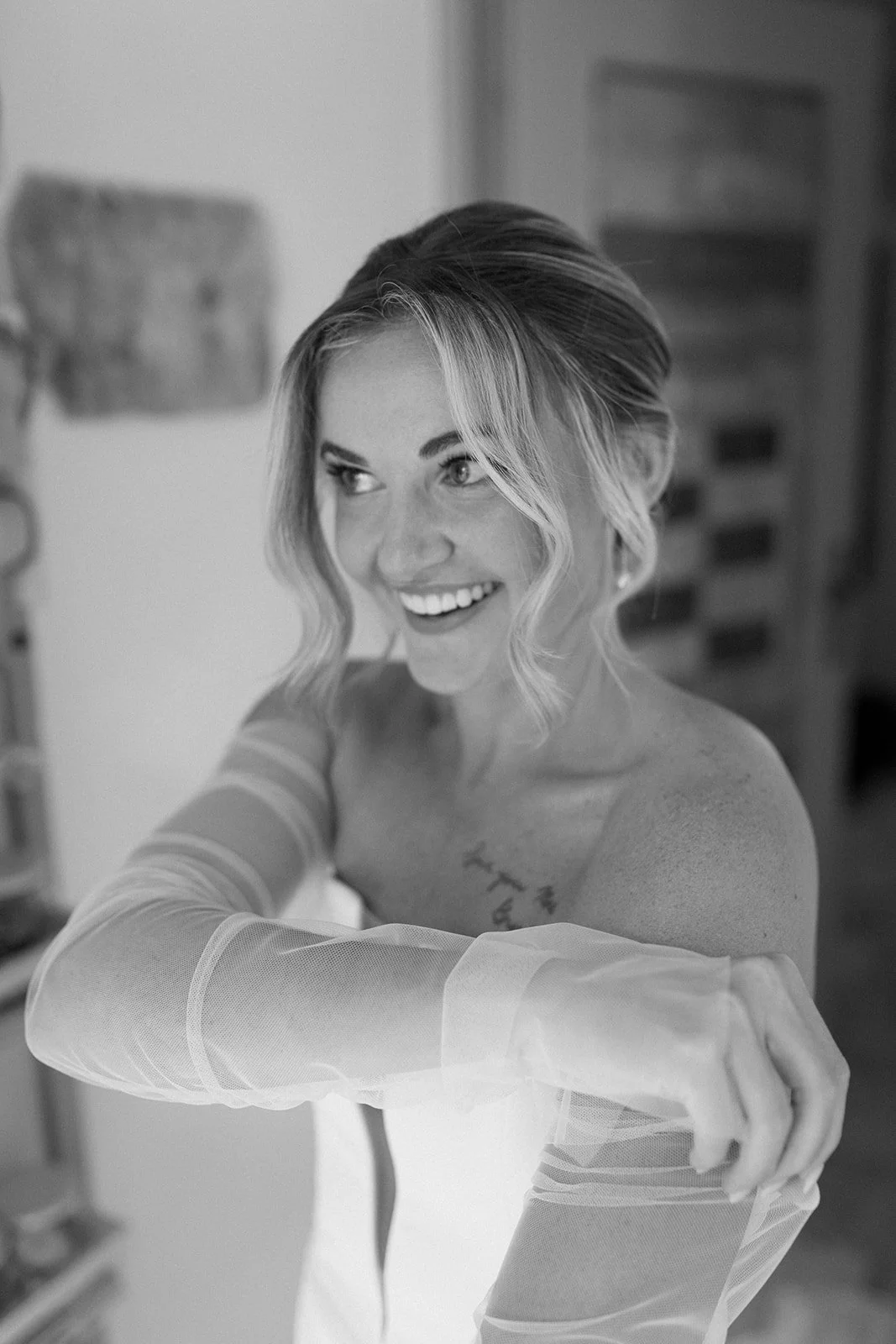A smiling woman with wavy hair and a tattoo on her chest, wearing light-colored sheer sleeves, posing indoors.