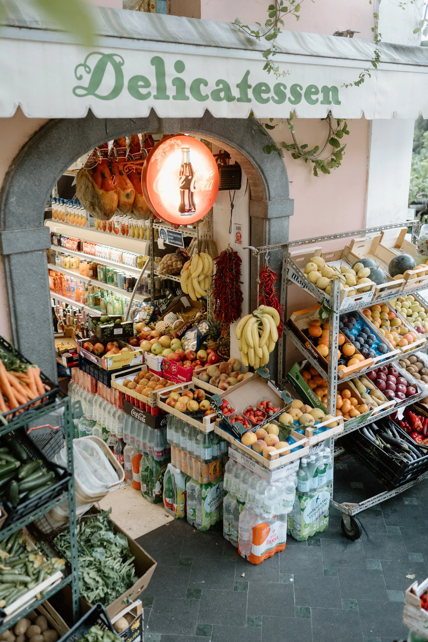 A small outdoor produce stand with a sign that reads 'Delicatessen' in green letters on a white awning. The stand displays various fresh fruits, including bananas, peaches, apples, and watermelons, along with shelves stocked with bottles of beverages