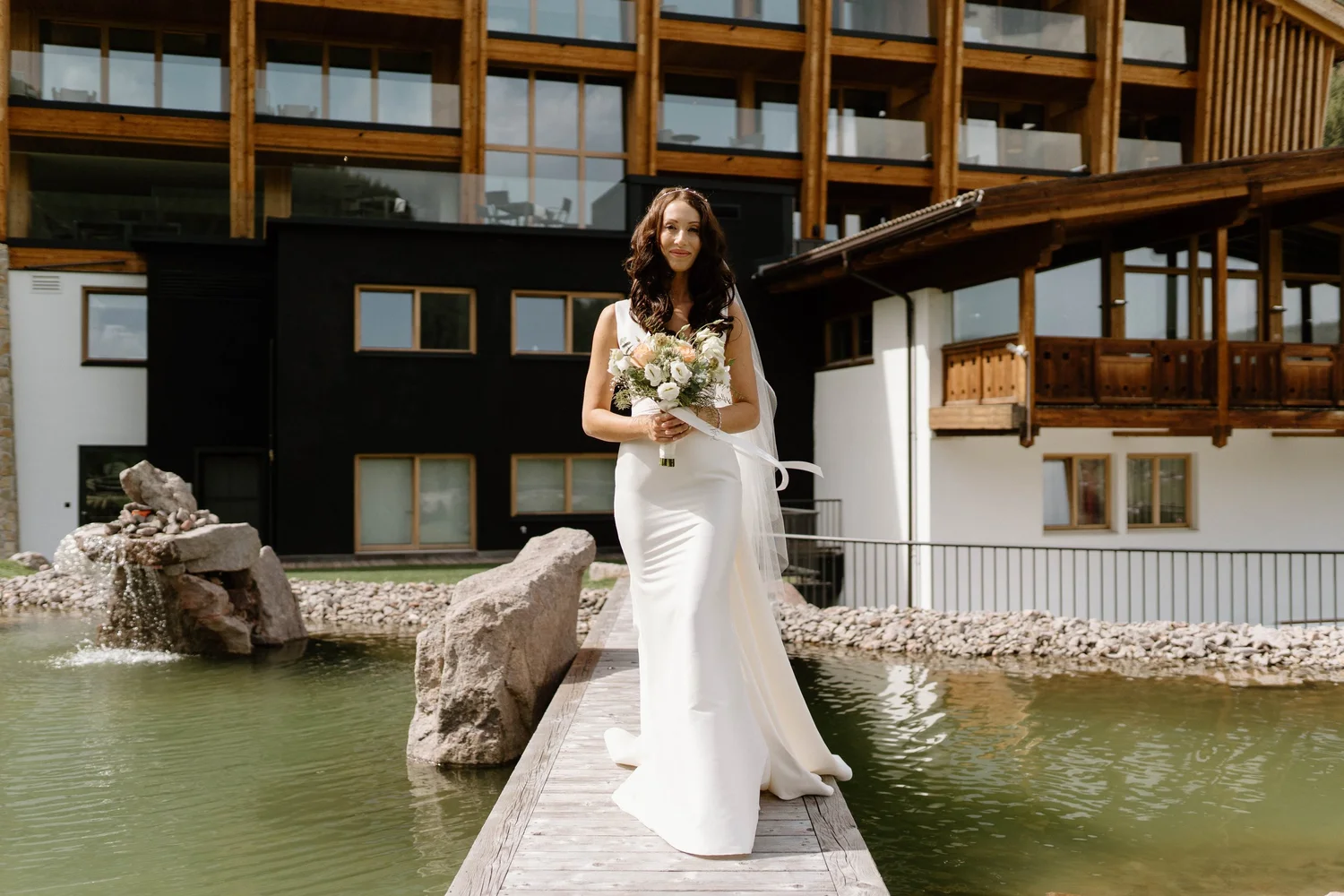 A bride in a white wedding dress holding a bouquet of flowers standing on a wooden dock over a pond with rocks and a fountain, with a modern multi-story wooden building in the background.