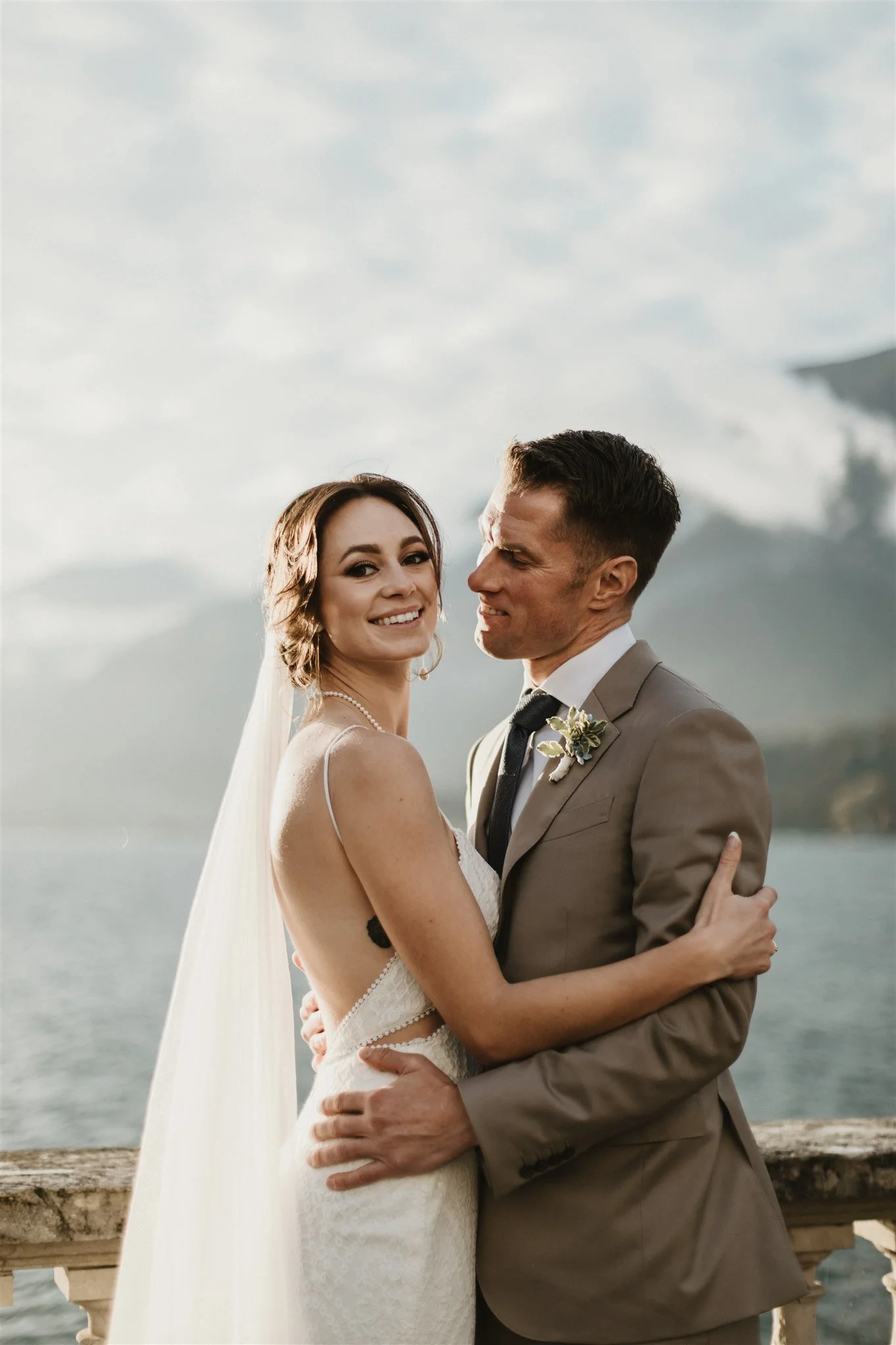 A bride and groom pose together outdoors near water, with the bride wearing a white lace wedding dress and veil, and the groom in a tan suit with a dark tie and boutonniere, smiling and embracing each other.