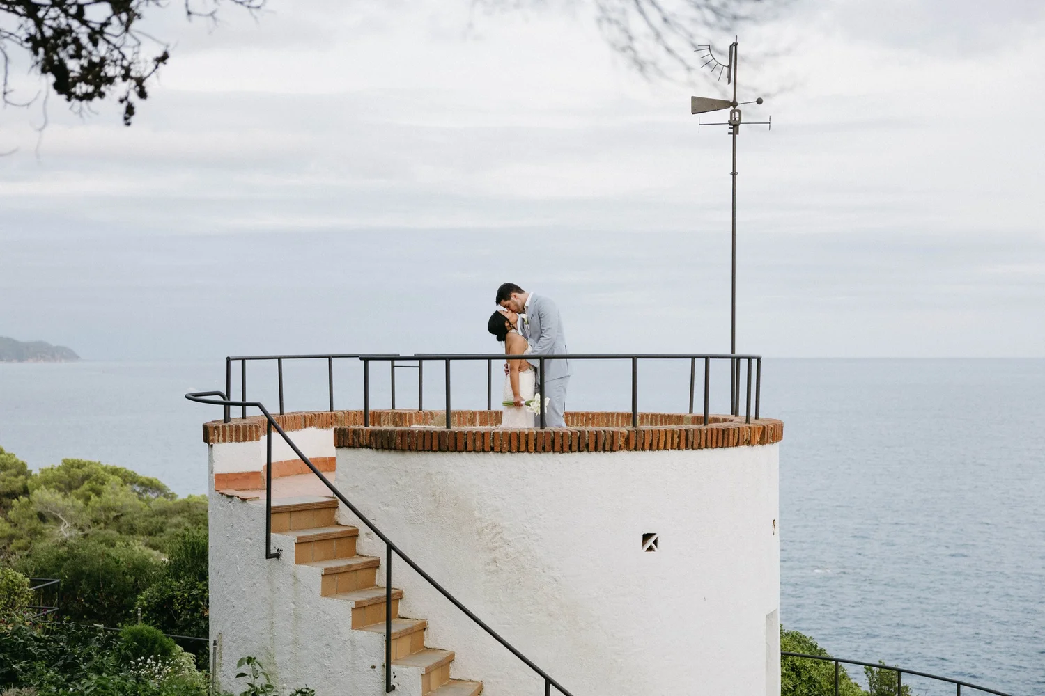 A couple in wedding attire sharing a kiss on a small brick and white stucco balcony overlooking the water, with an old weather vane on a pole and lush green trees in the background.