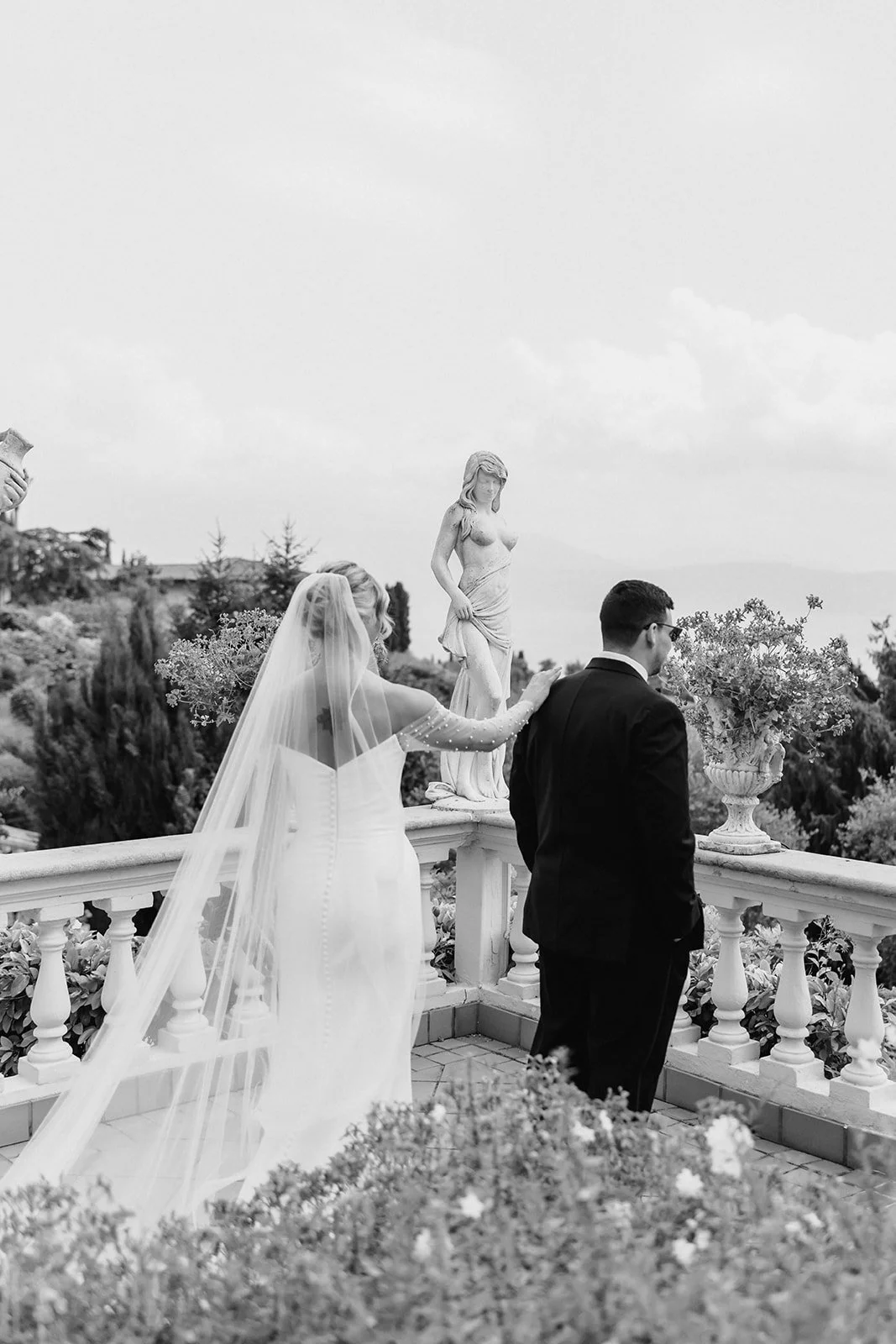 Bride and groom standing on a balcony with classical sculptures and potted plants, wedding ceremony in progress.