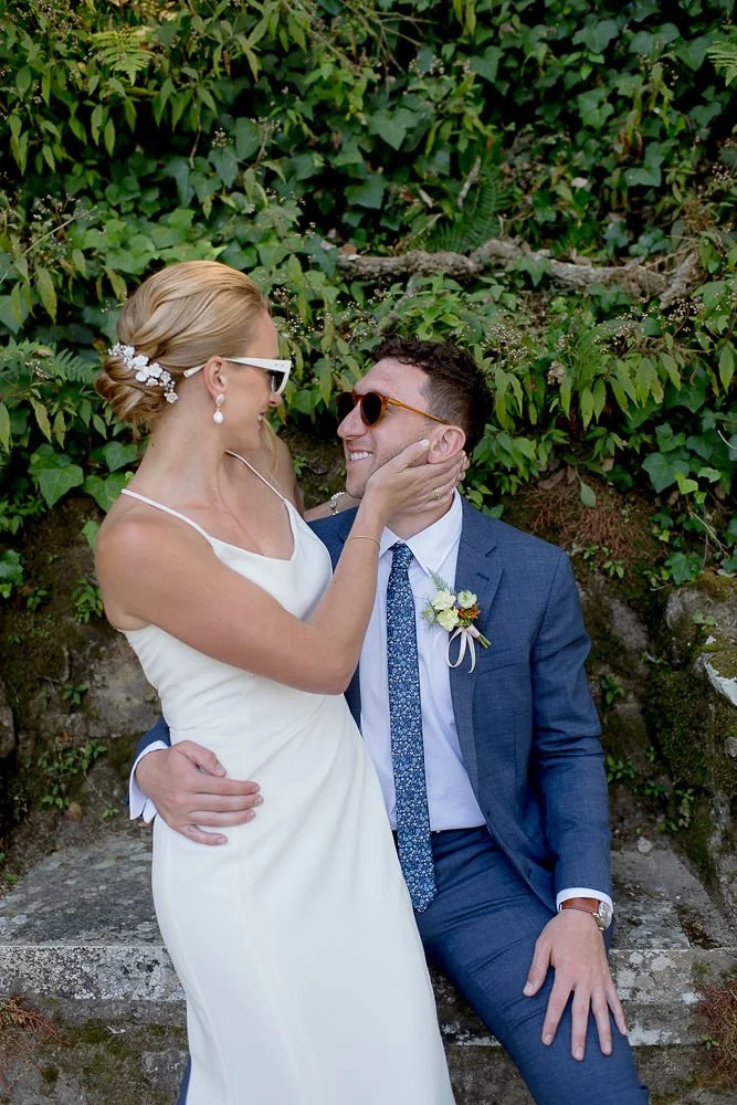 A smiling woman in a white dress and a man in a blue suit with a boutonniere sharing a joyful moment outdoors in front of greenery.