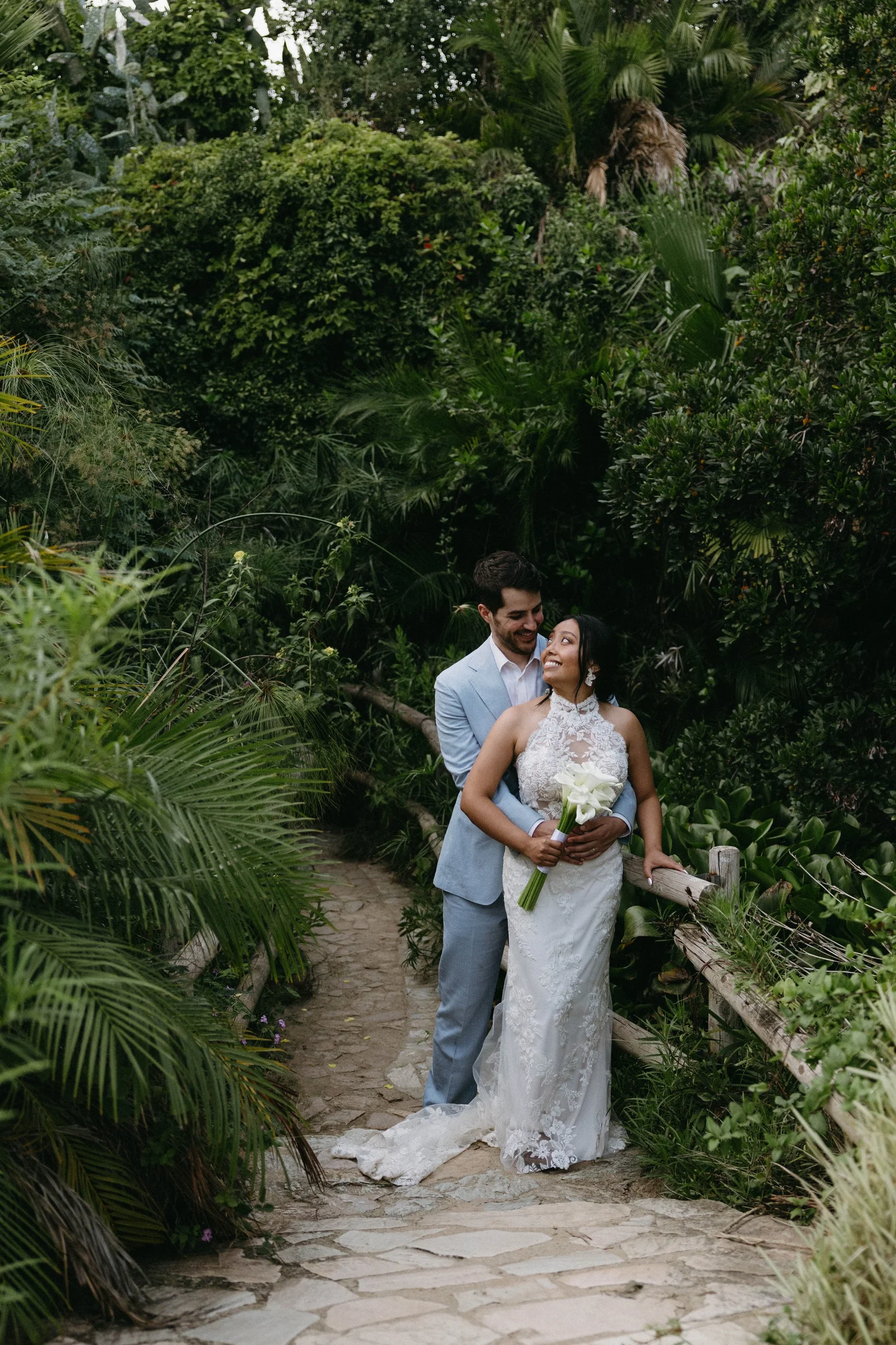 A couple in wedding attire standing on a stone pathway surrounded by lush greenery, holding hands and smiling at each other.