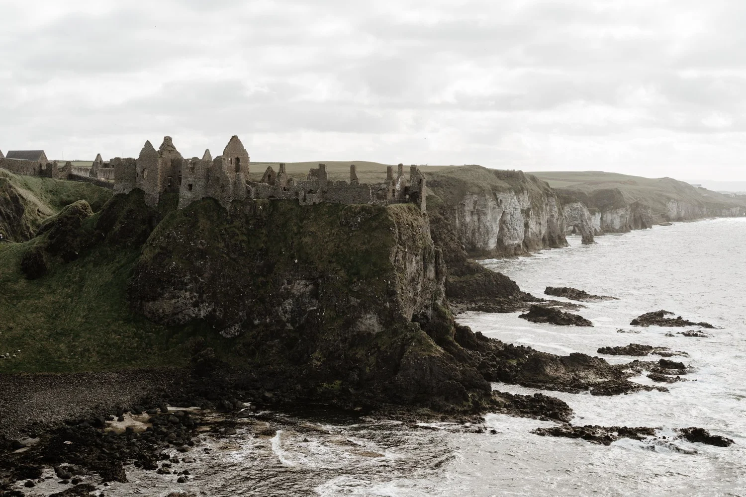 A photo of ancient stone ruins on a grassy cliff overlooking the ocean with rocky shoreline and cliffs in the background under a cloudy sky.