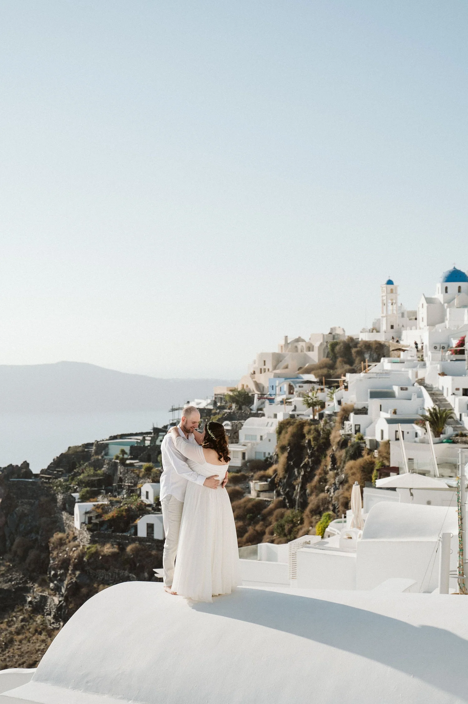 A couple sharing a dance on a white rooftop in Santorini, Greece, with white buildings and blue-domed churches in the background.