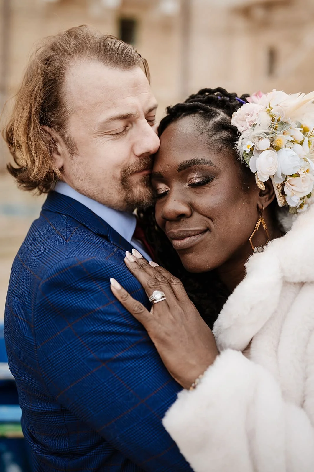 A couple embracing each other closely, with eyes closed, on a wedding day. The woman wears a floral headpiece and earrings, and the man wears a blue checked suit.