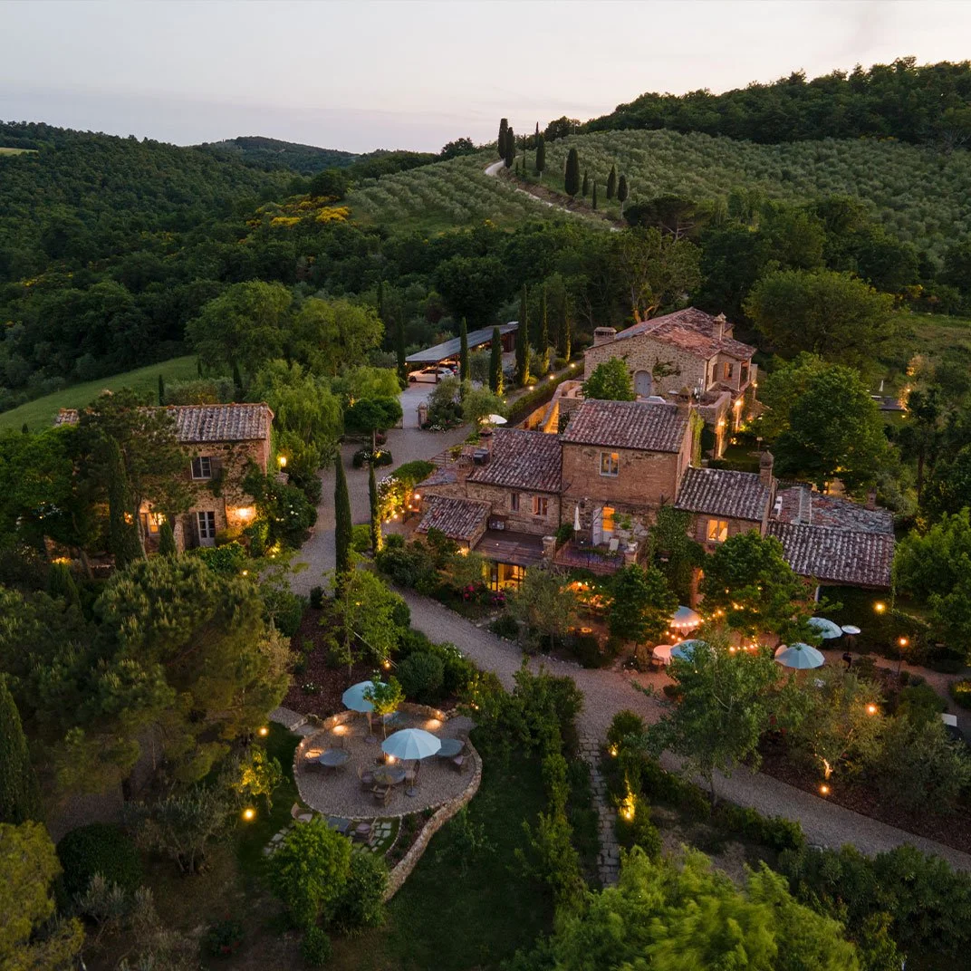 A scenic hillside estate with rustic stone houses, illuminated by warm outdoor lighting, surrounded by lush greenery, with a small patio area and winding pathways, set against rolling hills and trees in the background.