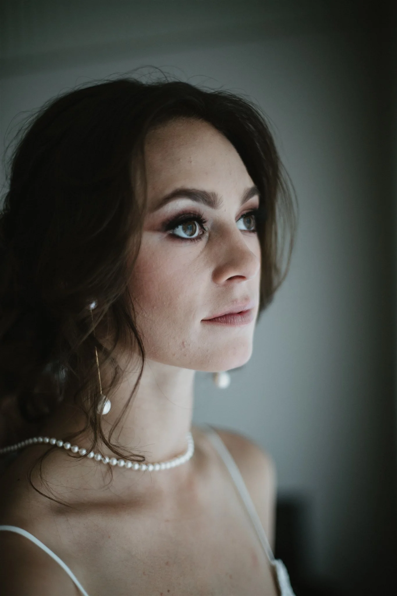 Close-up of a woman with dark wavy hair, wearing pearl jewelry, with makeup, looking to the side, in a soft focus.