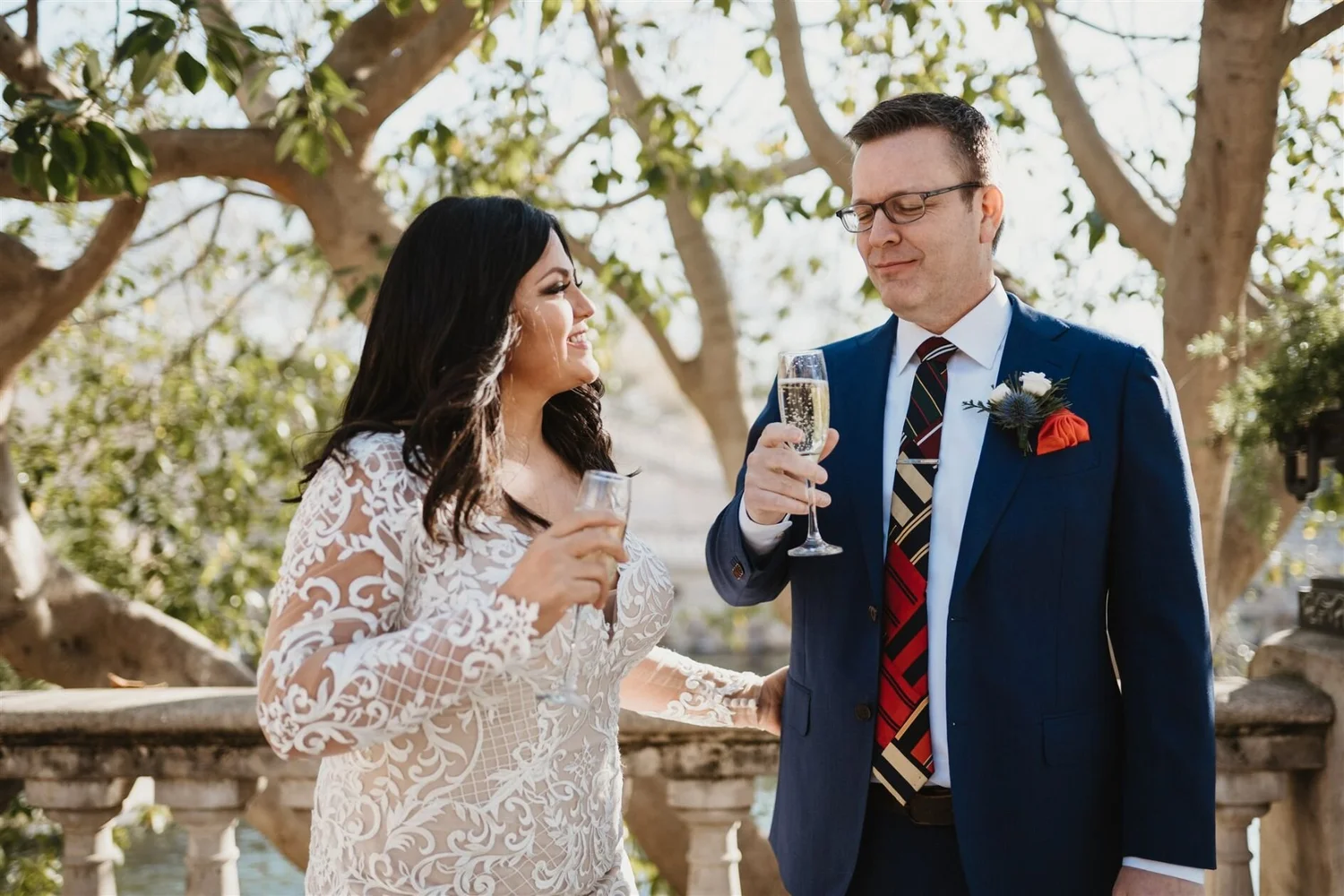 A bride and groom standing outdoors under a tree, holding champagne glasses, celebrating their wedding. The bride is in a white lace wedding dress and the groom is in a dark blue suit with a striped tie and boutonniere.