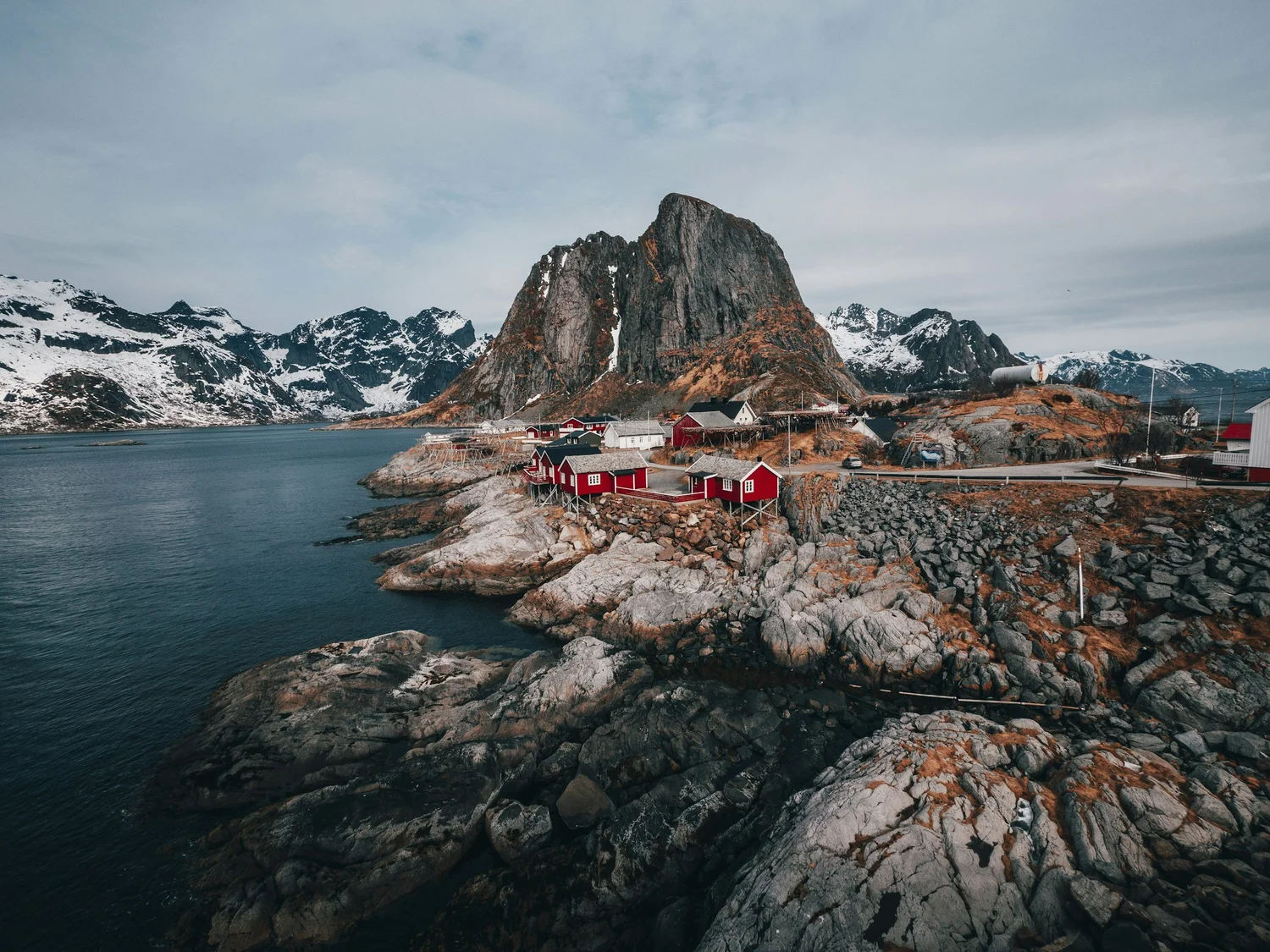 A large rocky mountain with snow patches overlooks a small coastal village with red and white houses, near a body of water under a cloudy sky.