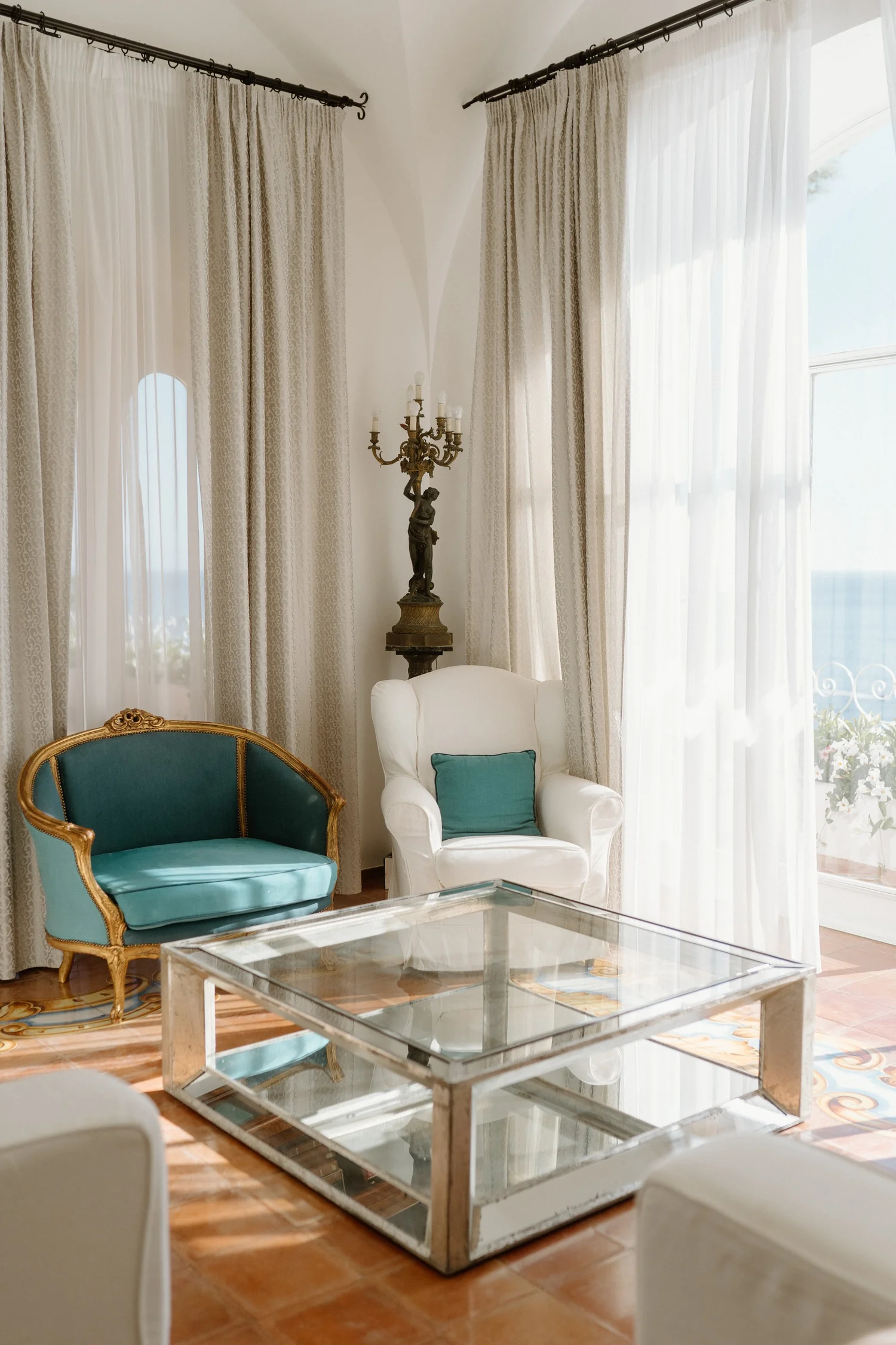 Living room with flowy white curtains, a vintage green and gold armchair, a white armchair with a teal pillow, an ornate antique chandelier on a pedestal, and a glass coffee table with a white frame.