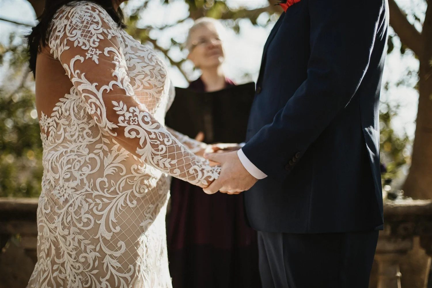 A bride and groom holding hands during their wedding ceremony outdoors, with a woman officiant in the background.