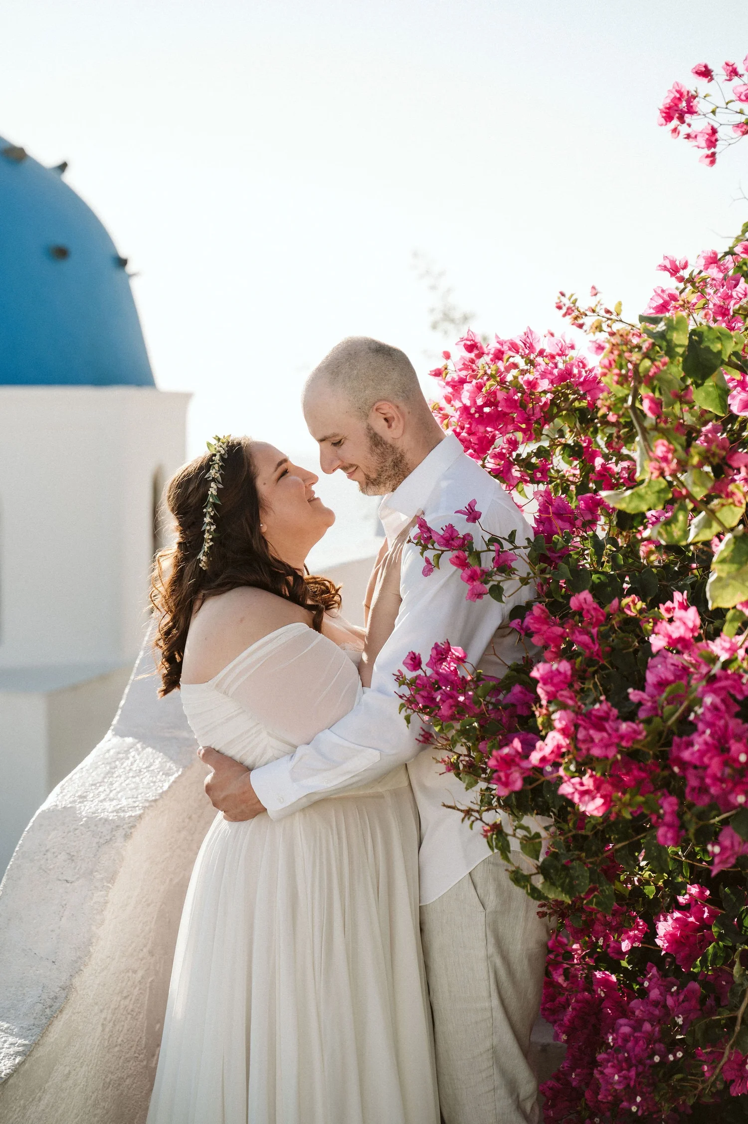 A couple dressed in wedding attire embracing outdoors near vibrant pink bougainvillaea flowers, with a white building and a blue-domed church in the background, typical of Santorini, Greece.