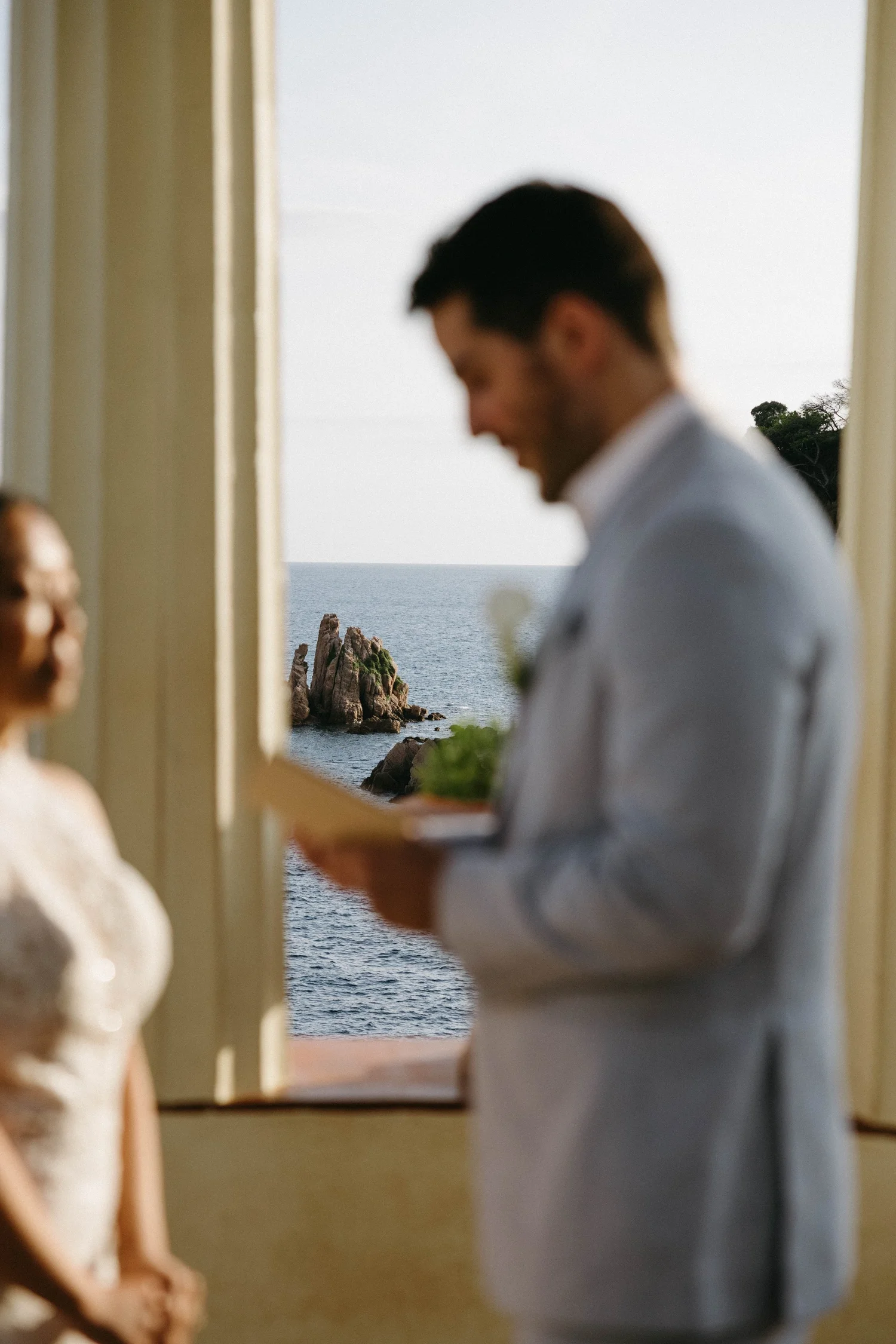 A man in a white shirt is holding a book or a folder and smiling, standing by an open window with a view of the ocean and rocky cliffs.