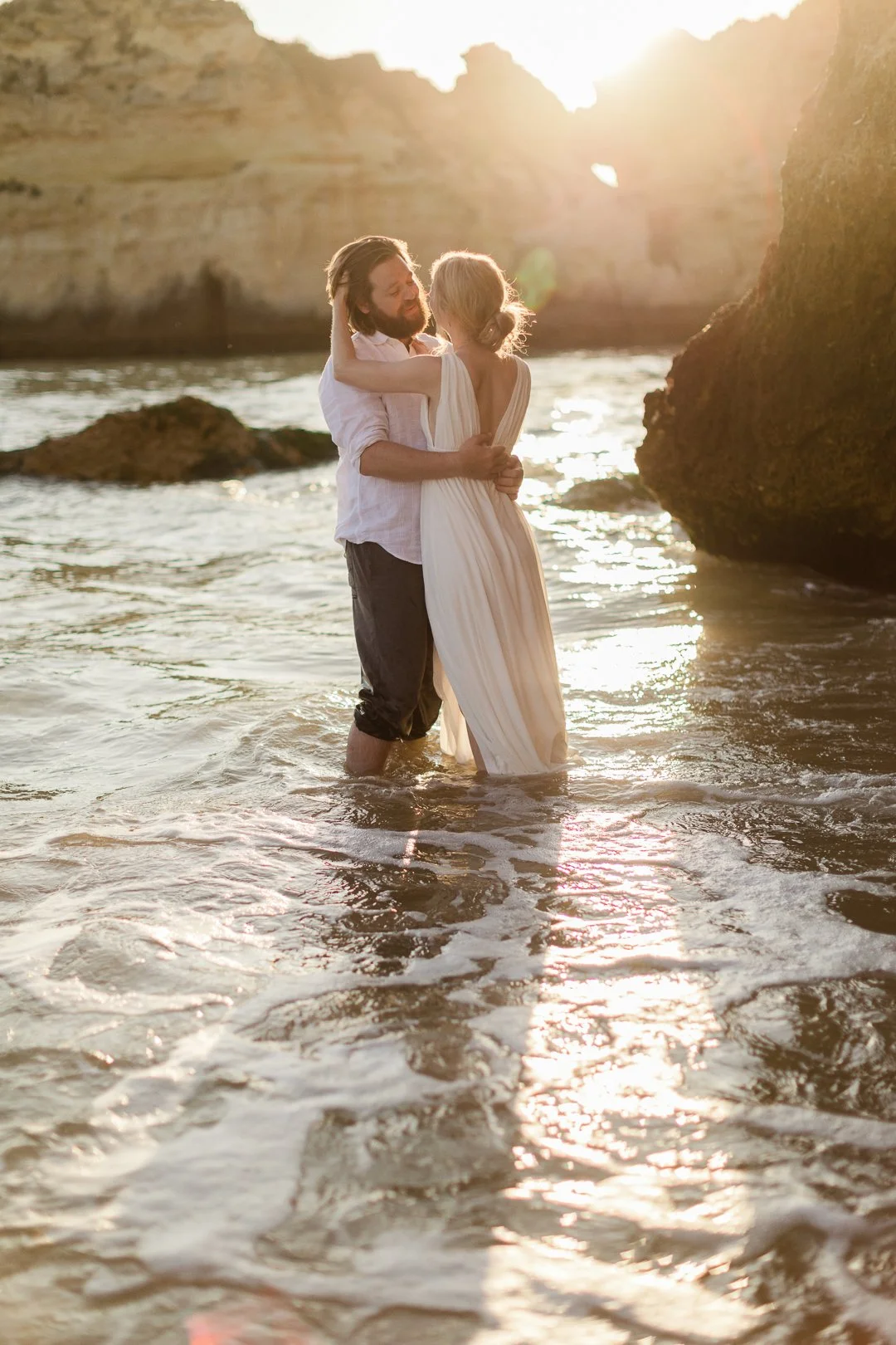 A couple stands in the shallow water of a beach at sunset, embracing each other. The man wears a white shirt and dark pants, and the woman wears a flowing white dress. Large rocks and cliffs are visible in the background with the sun setting behind them.