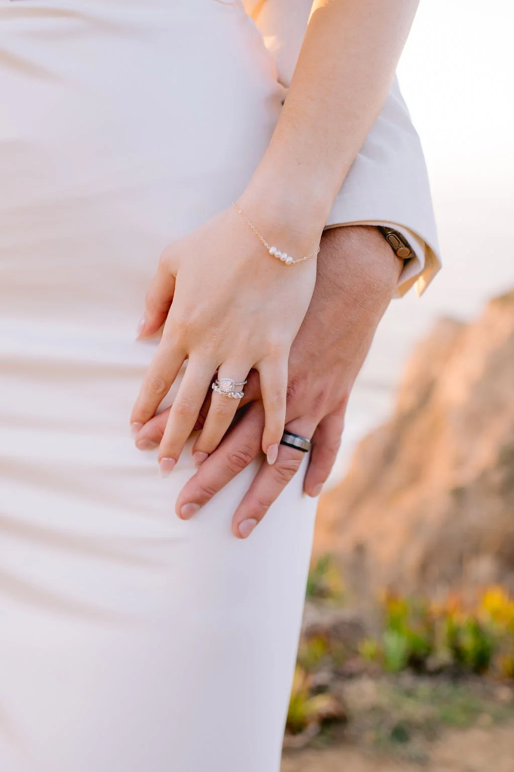 Close-up of a couple's hands gently holding each other, showing wedding rings and a bracelet, with a blurry outdoor background.