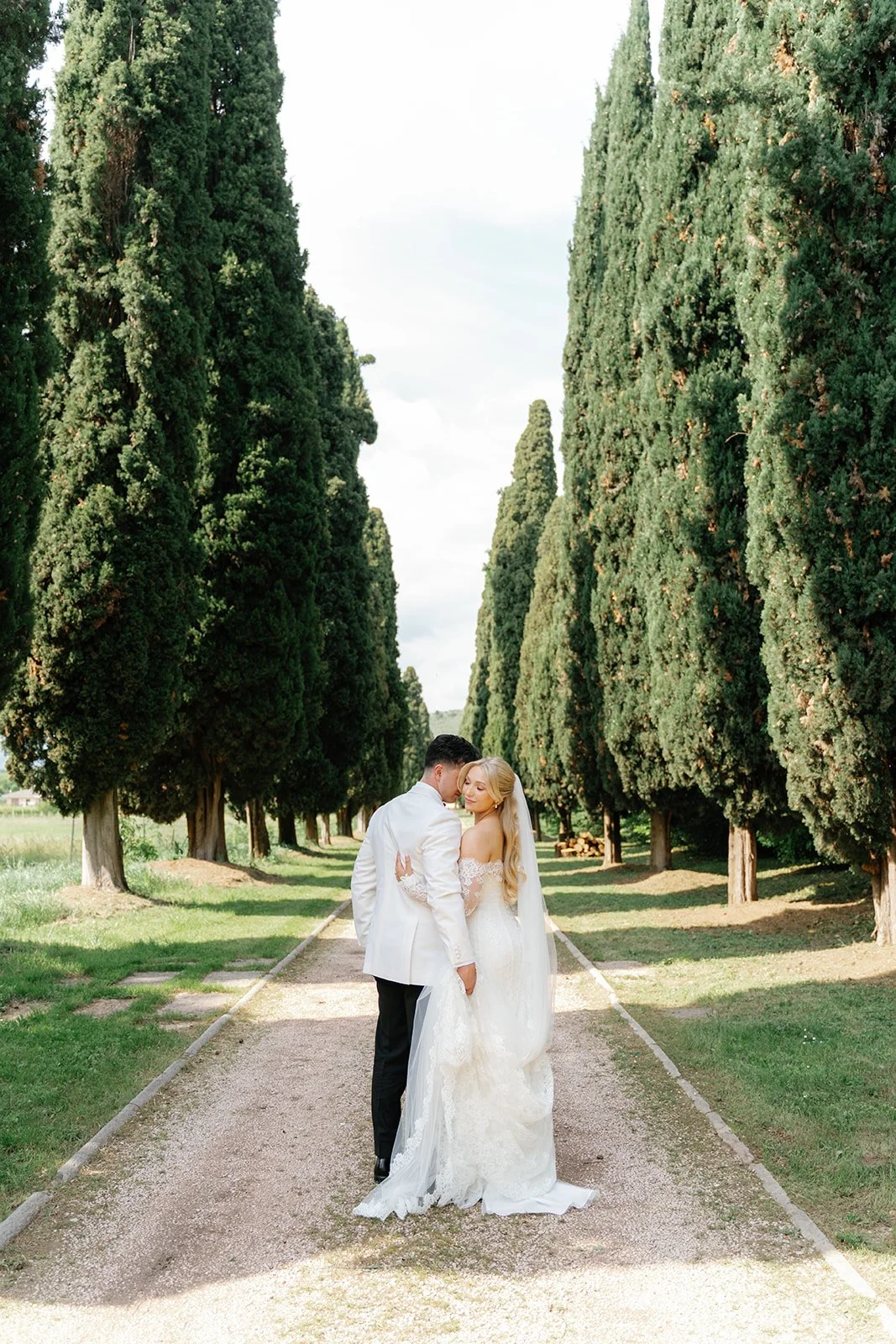 A bride and groom sharing a moment on a tree-lined pathway, surrounded by tall, lush green trees.