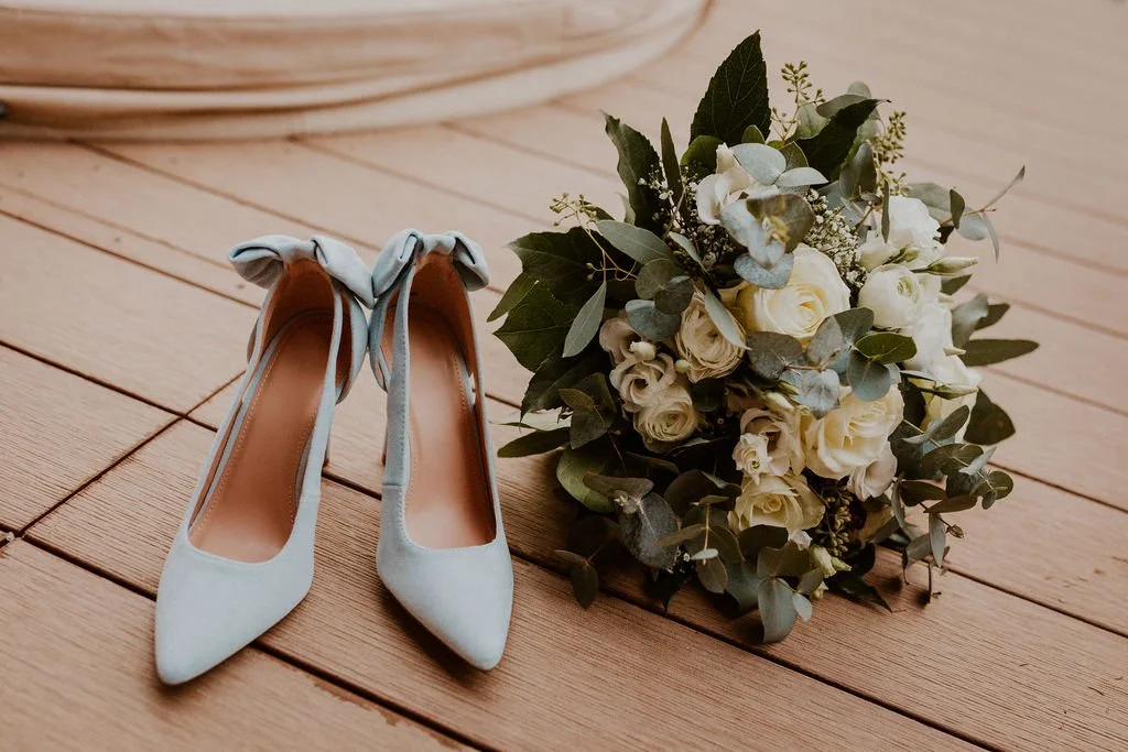 A pair of light blue high heels with bows on the back, placed next to a bouquet of white flowers and green leaves on a wooden floor.