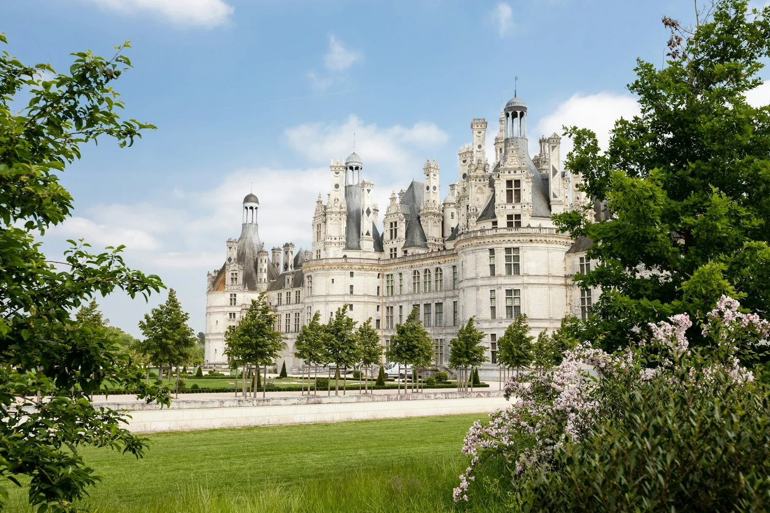 A large, historic castle surrounded by lush green trees and a well-manicured lawn, with a partly cloudy sky in the background.