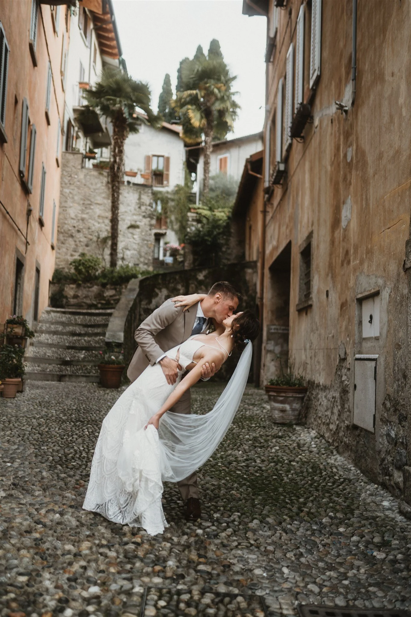 A newlywed couple sharing a kiss in a narrow, cobblestone alley with tall, old buildings, palm trees, and potted plants in the background.