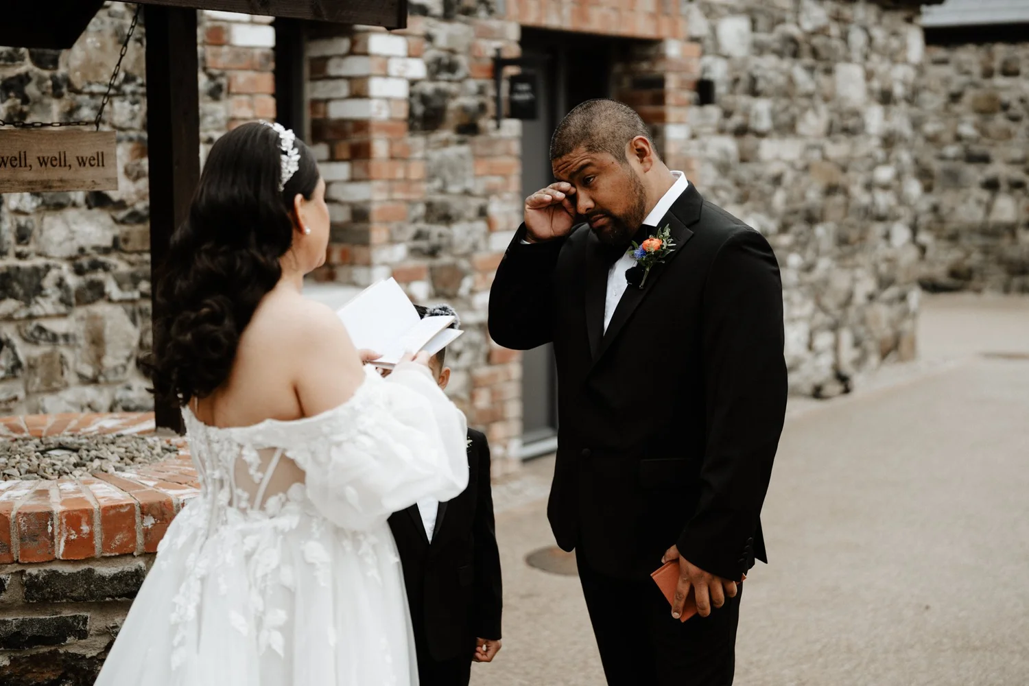 A bride and groom stand outside during a wedding ceremony. The bride, dressed in a white lace gown with off-the-shoulder sleeves, is reading from a book, while the groom, in a black tuxedo with a colorful boutonniere, wipes a tear from his eye. A you