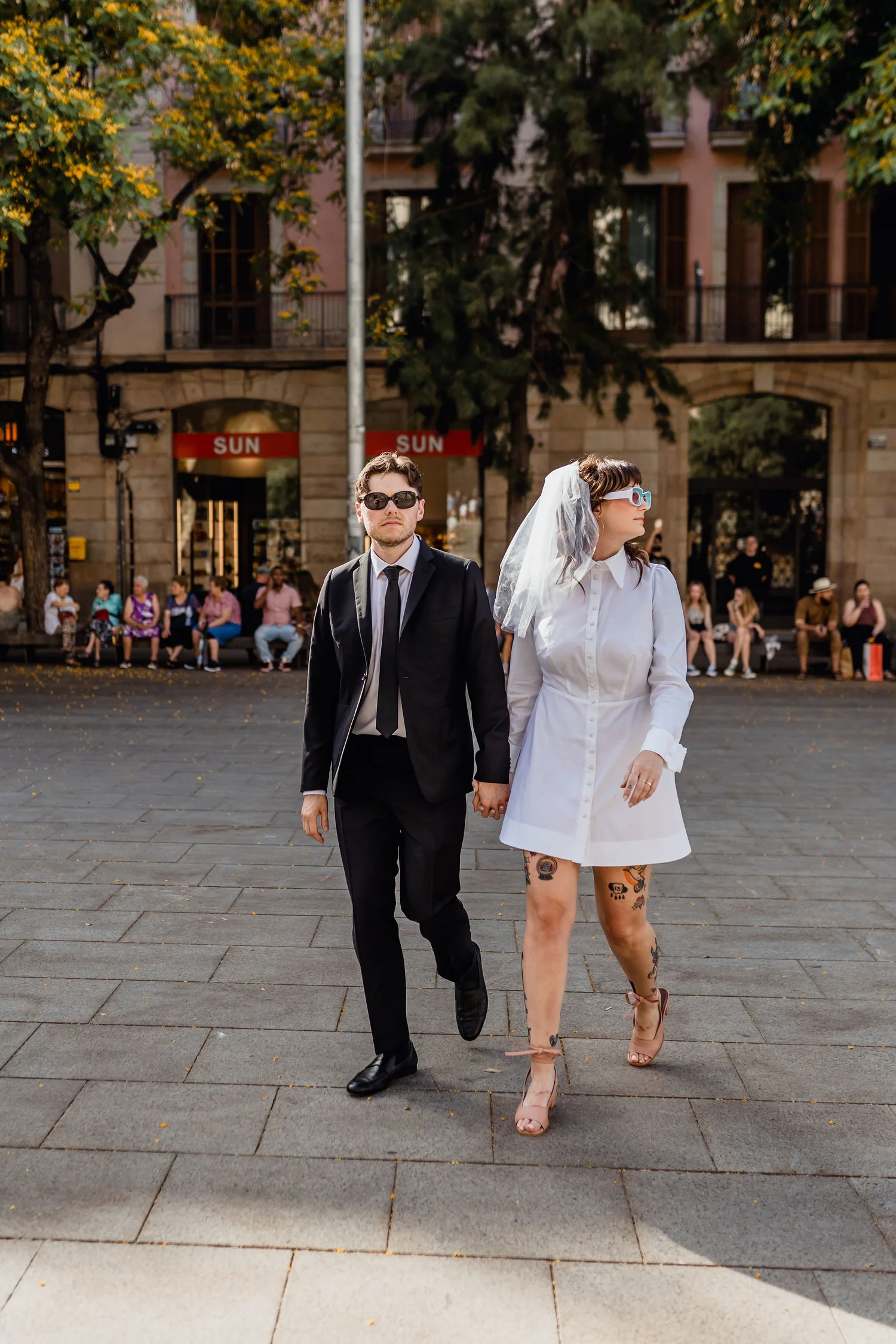 A man and a woman hold hands while walking on a city street, with people sitting on a bench in the background and colorful trees lining the street.