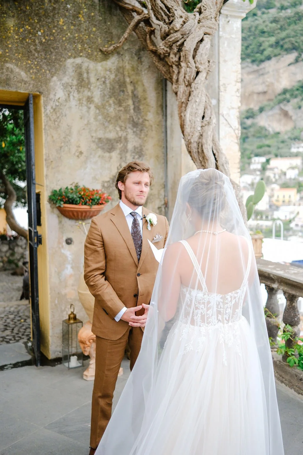 A bride and groom during their wedding ceremony outdoors on a terrace with a scenic hillside view in the background. The groom is wearing a brown suit, and the bride is dressed in a white gown with a veil.